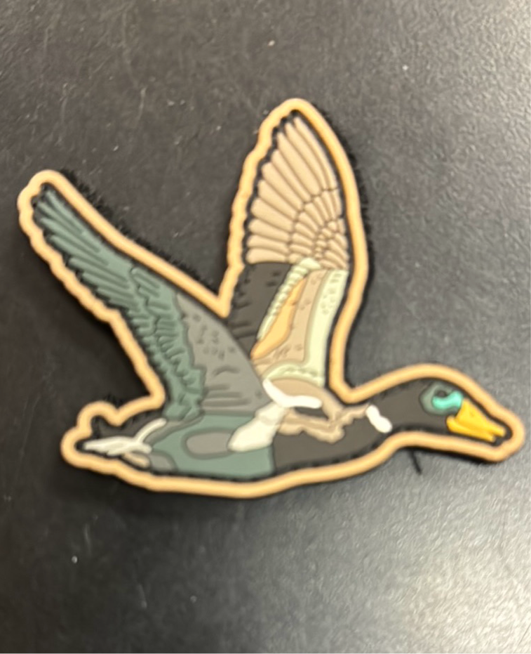 The OG Duck It PVC Patch by Mucker Bucket showcases an illustration of a duck flying with detailed feathers in green, brown, and beige tones, a yellow beak against a dark background, and fully extended wings.