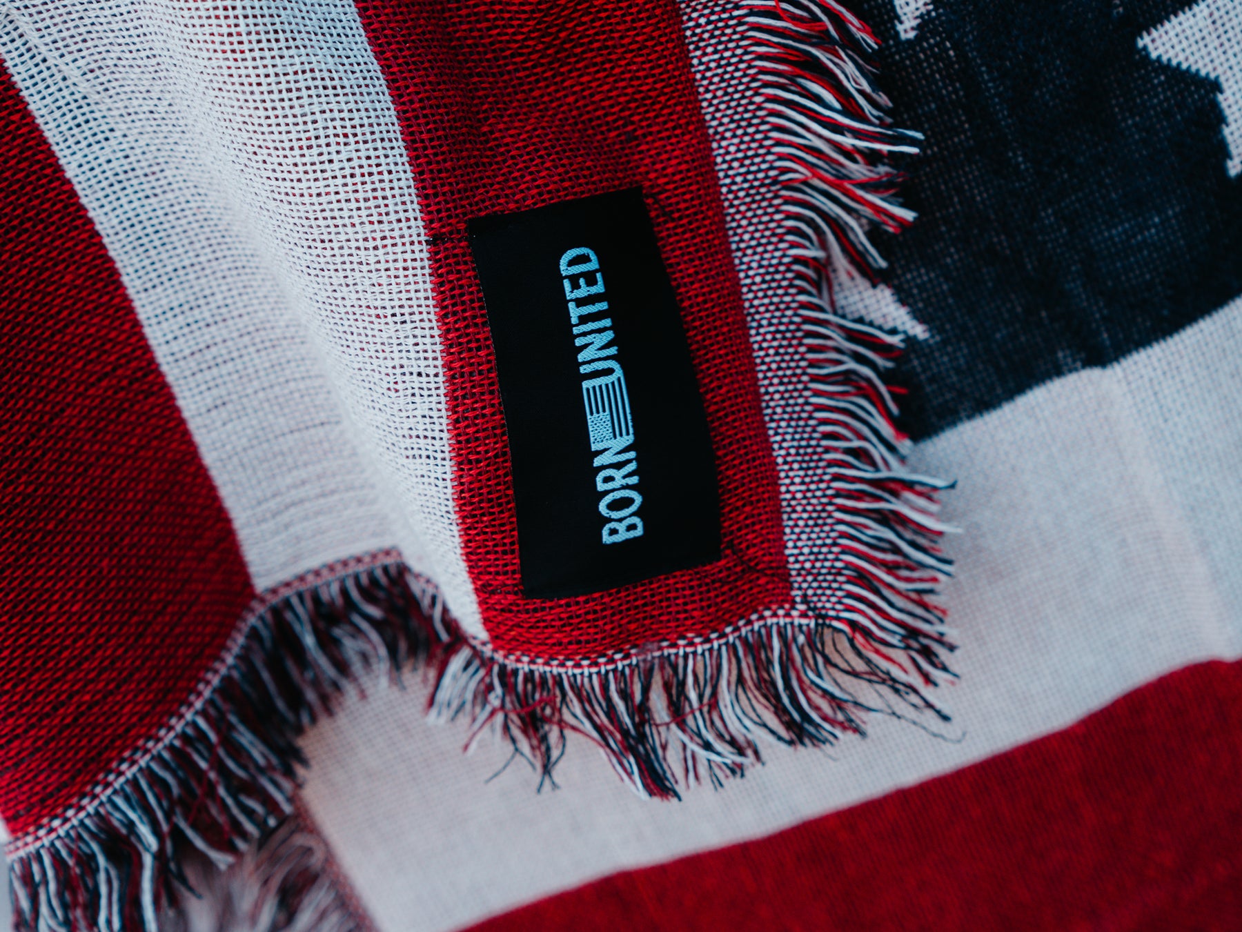Close-up of the American Flag Jacquard Blanket featuring red, white, and blue resembling Old Glory. A black tag reads BORN UNITED. The fringe adds texture to the edge. Brand: Born United Private Label.