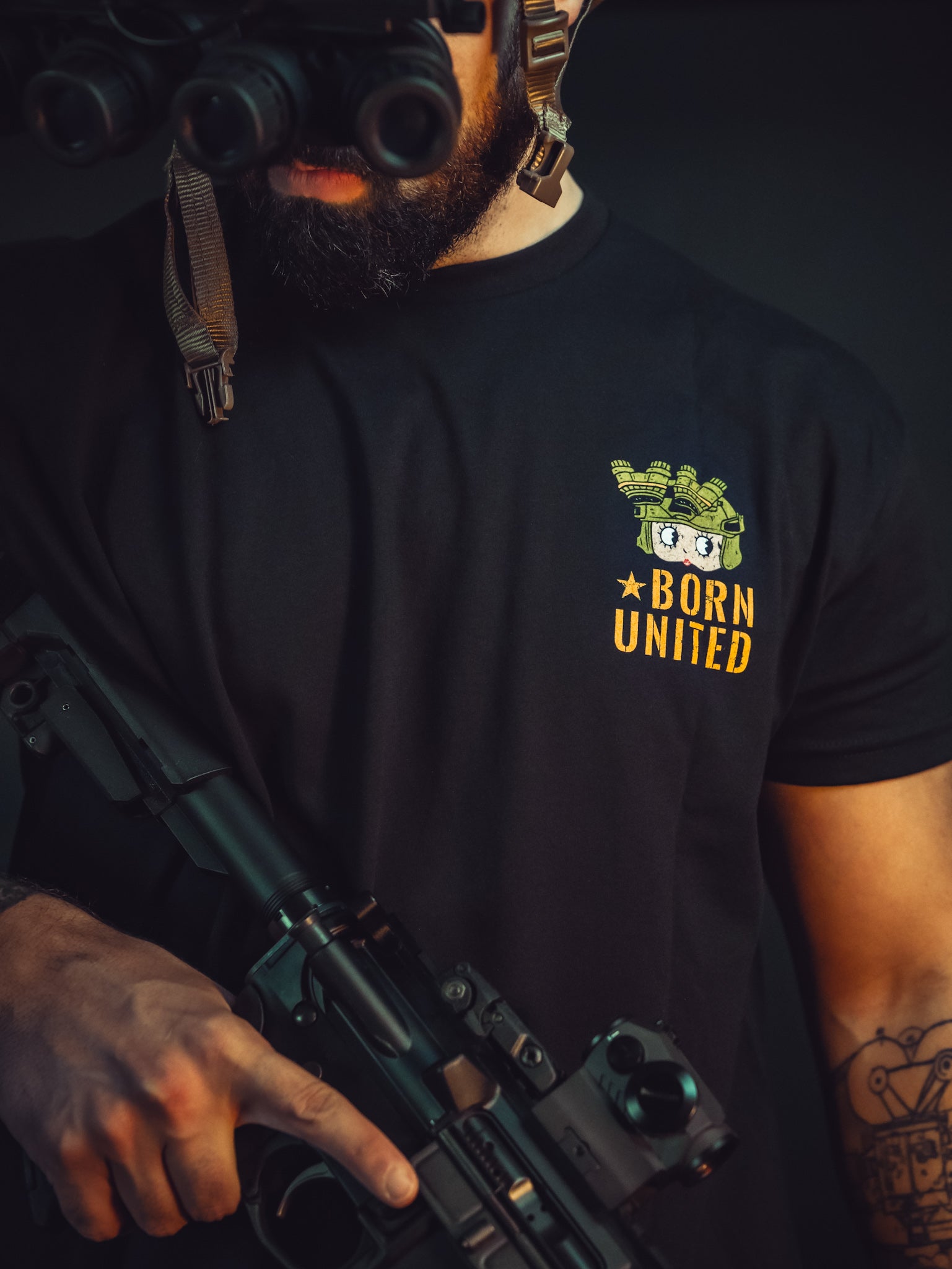 A bearded man in a black Betty Boop Tee by Born United Private Label, symbolizing freedom, grips a rifle. He peers through night vision goggles, tattoos visible on his arm. Defenders like him stand vigilant in the dark background.