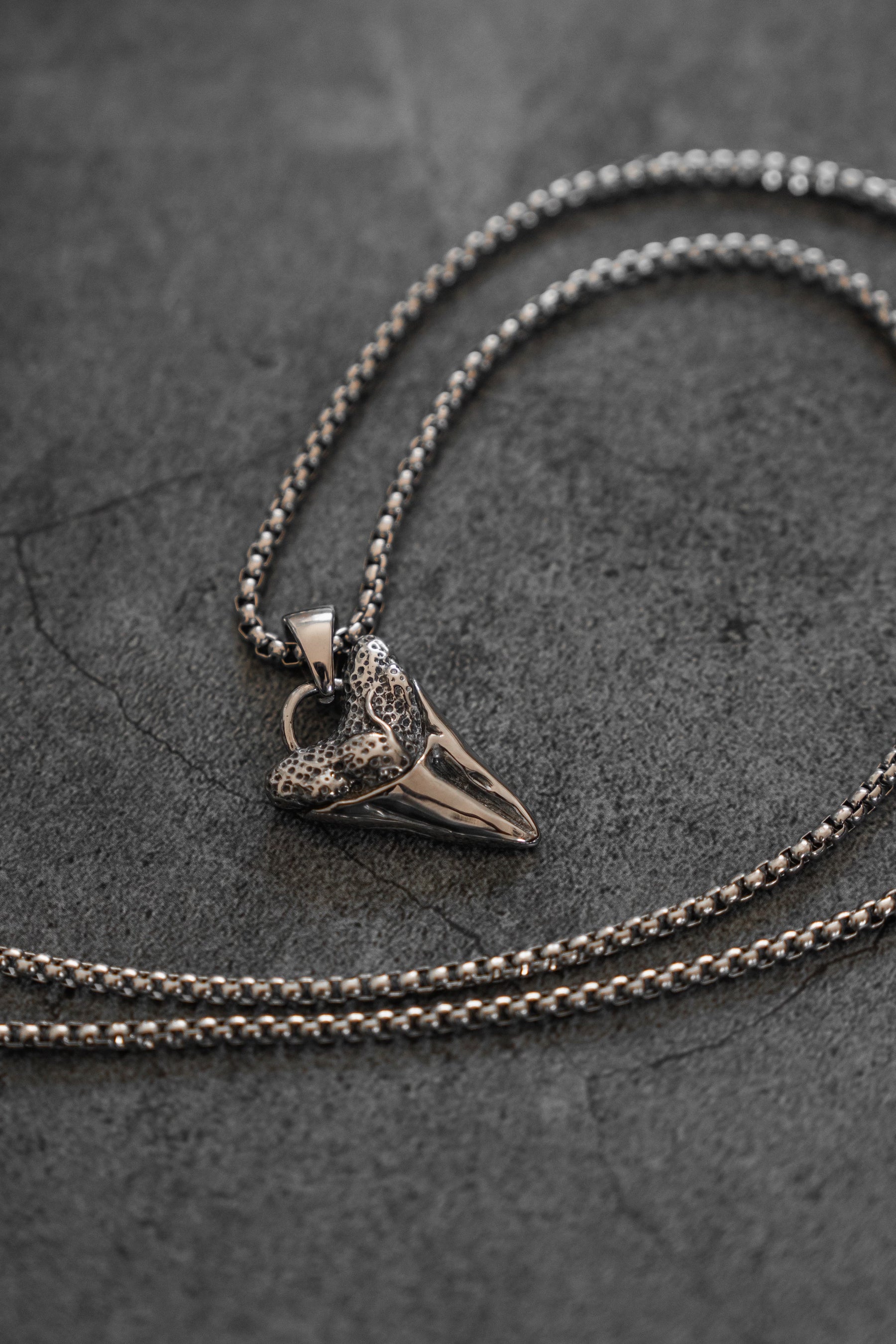 The Relentless Predator Necklace by Relentless Betrayal showcases a stainless steel pendant shaped like a detailed, textured heart atop an arrowhead, resting on a dark, textured surface.