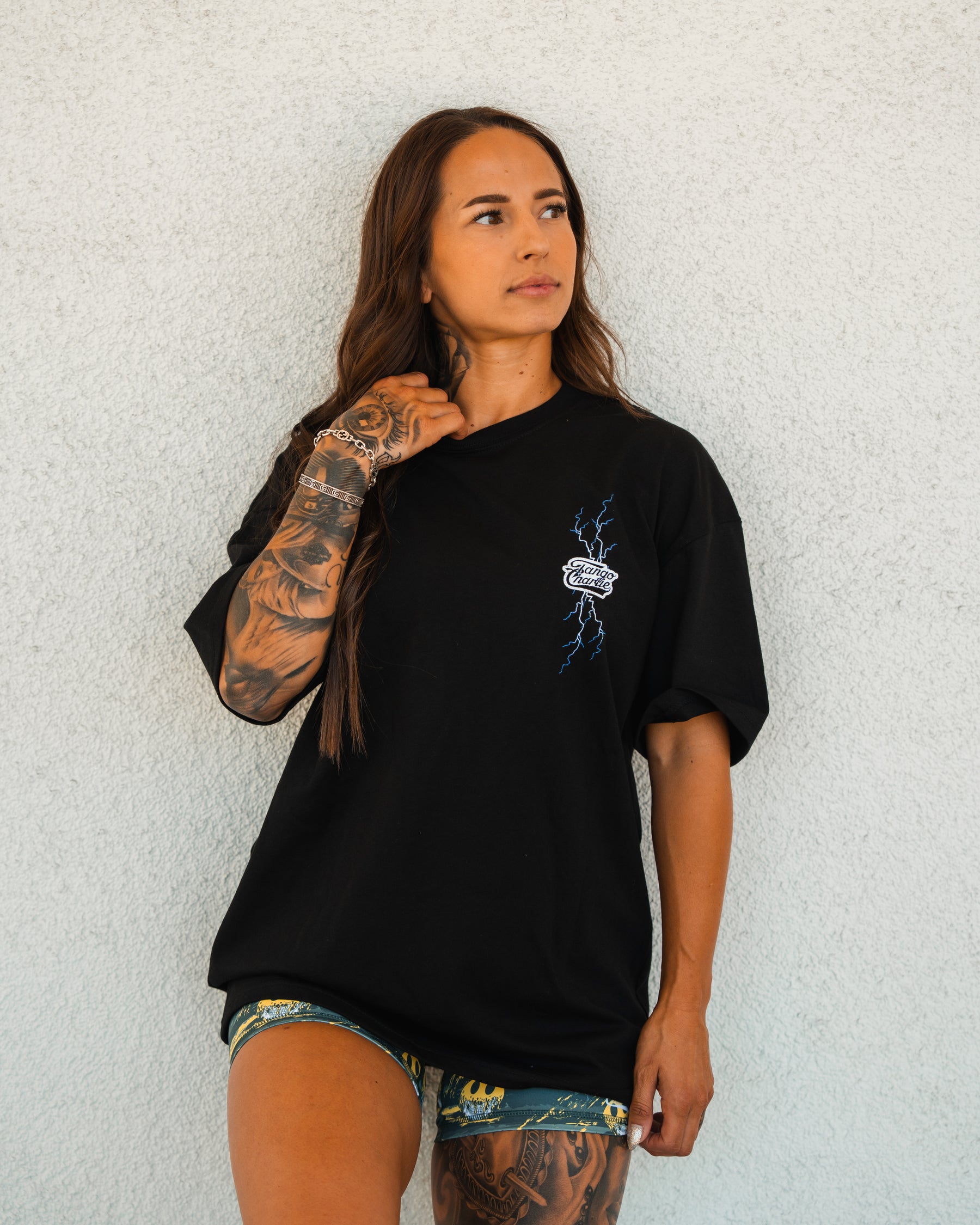 A woman with long brown hair and tattooed arms stands against a light wall, wearing the Tango Charlie Apparel Locked In Premium Oversized Tee in black with a small graphic, paired with patterned shorts, looking to the side.