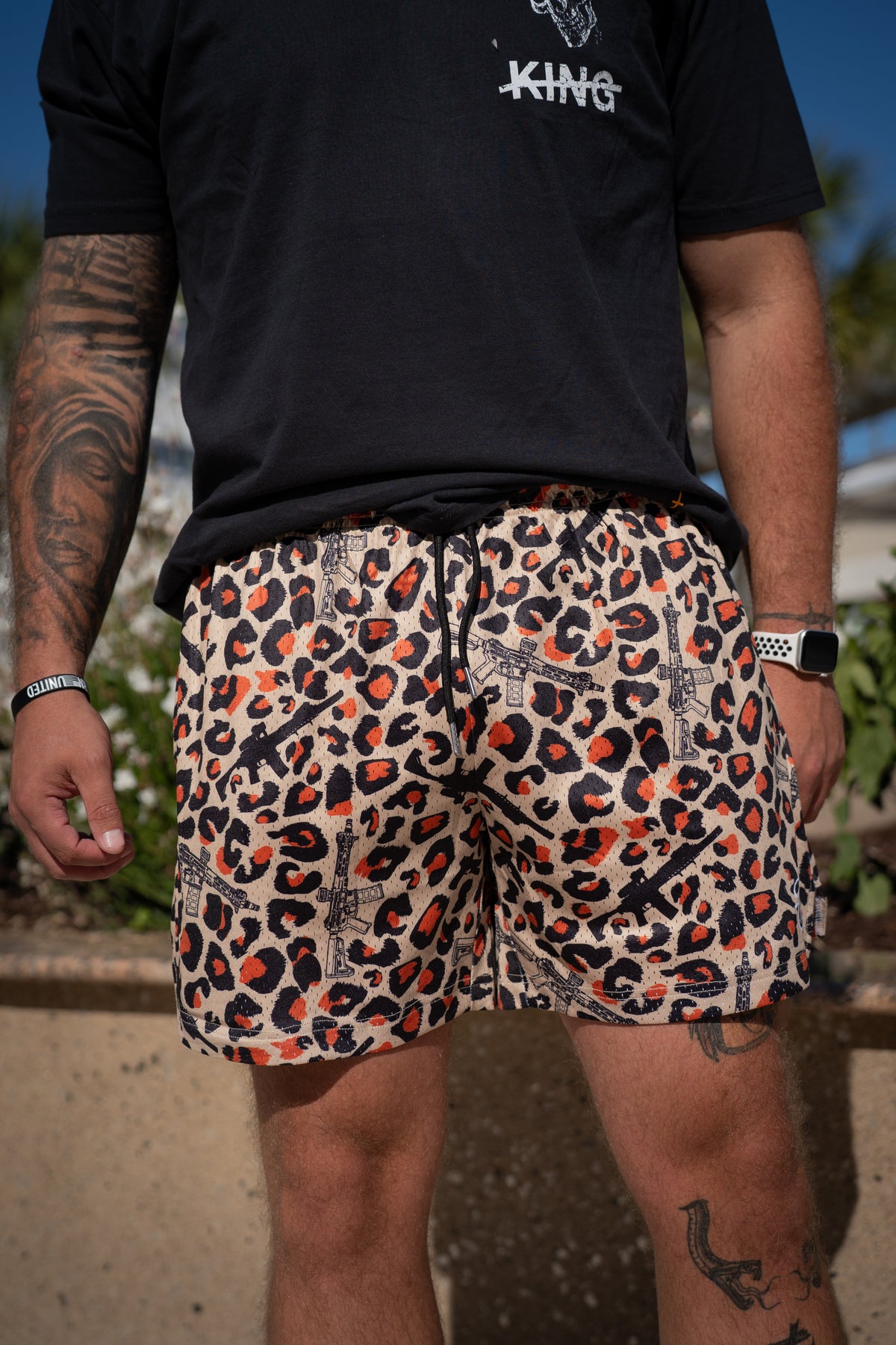 A person with visible arm and leg tattoos, a bracelet, and a ring stands outdoors among green plants and a stone wall, wearing Tango Charlie Apparel's Men's Mesh Lifestyle Shorts | 5.5"- Loaded Leopard and a black t-shirt.