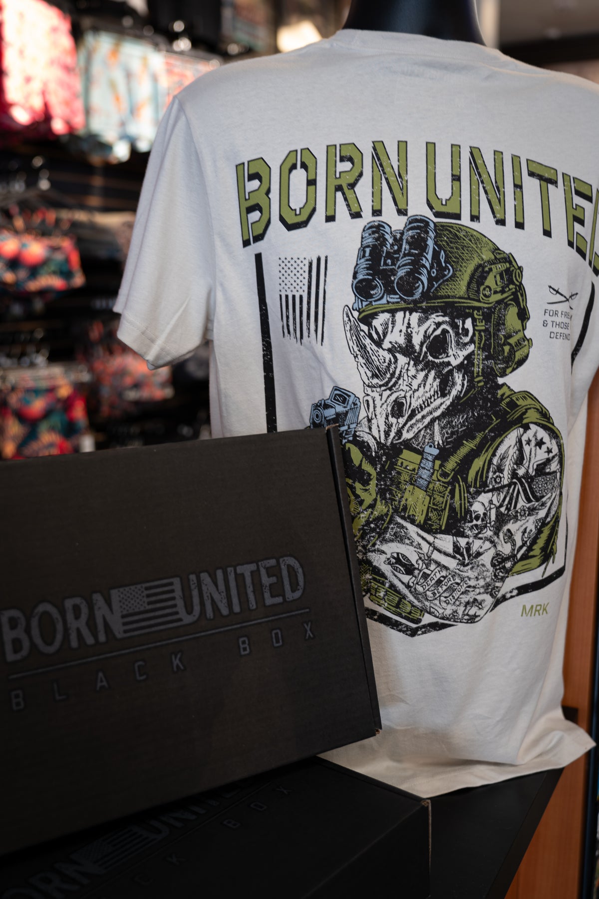 A mannequin wears a white Born United Private Label tee with a skull in military gear. In front, the Born United Black Box - TactiRhino from our Mystery Tees collection partially obscures the shirt.