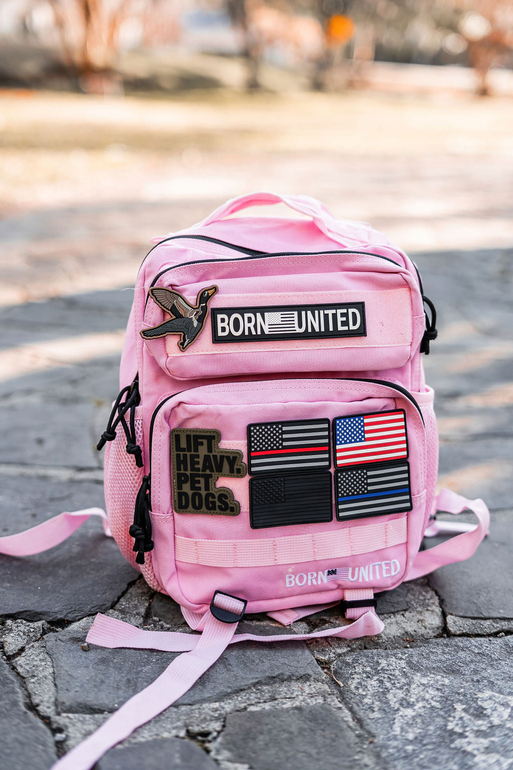 The Pink Mist Premium Tac-Pac - 15L from Born United Private Label sits on a stone surface, featuring patches saying BORN UNITED, multiple flags, and LIFT HEAVY. PET DOGS. It has several compartments with black zippers and straps.