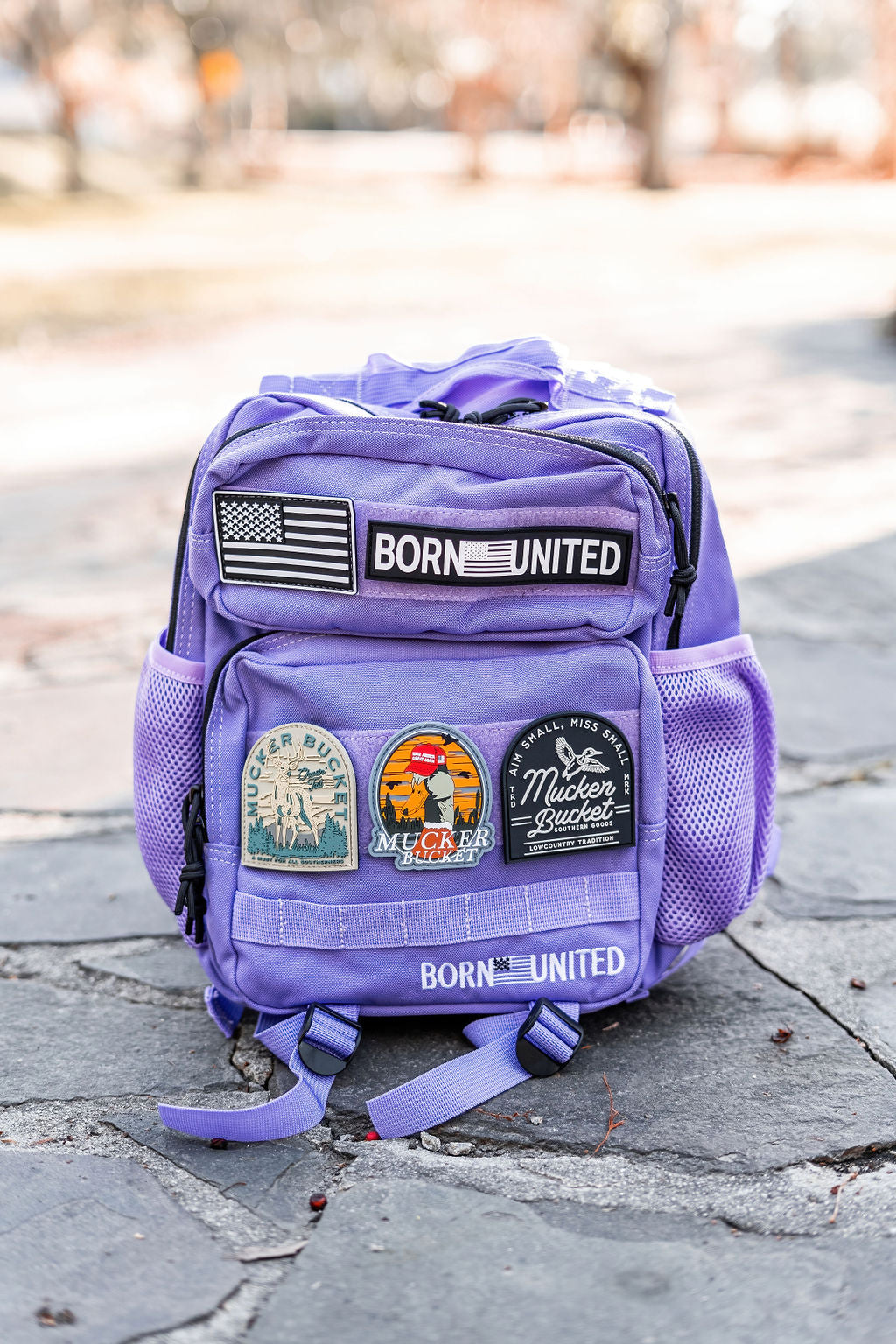 The Imperial Purple Tac-Pac - 15L from Born United Private Label is uniquely styled with multiple patches, including a U.S. flag and BORN UNITED on the top flap, and vibrant round patches on the front set against a picturesque stone path in a park setting.