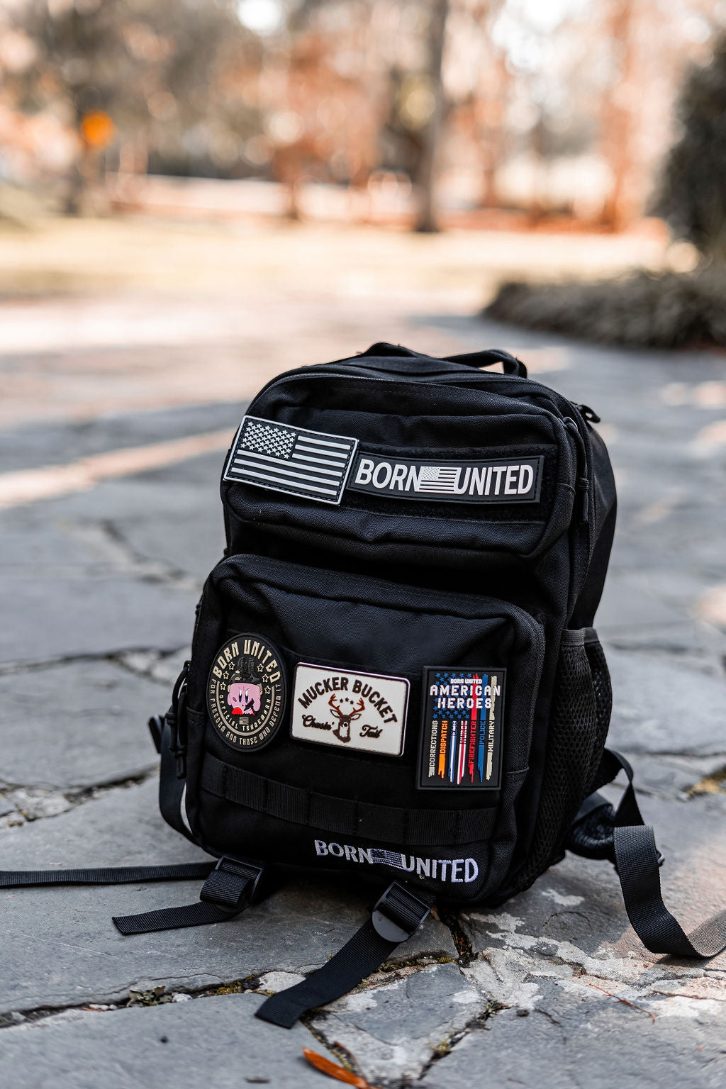 The Midnight Black Premium Tac-Pac - 15L by Born United Private Label rests on a stone path, adorned with patches like an American flag, BORN UNITED, and military-themed badges. The blurred garden background showcases green and brown tones, enhancing the scenes charm.