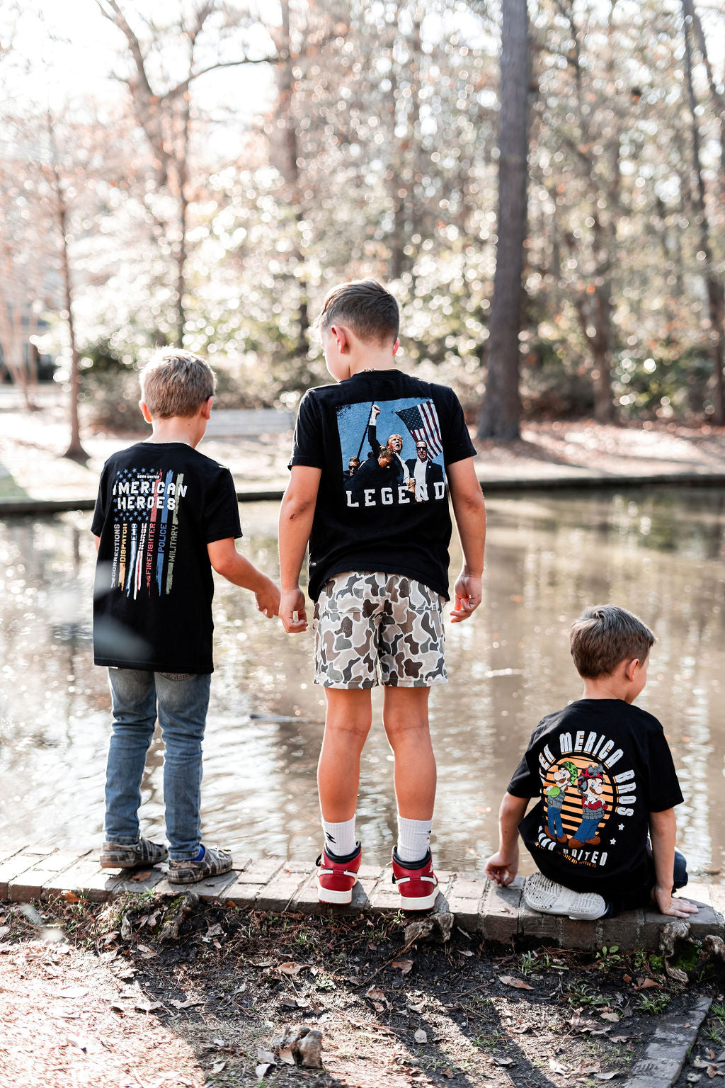 By a pond, three boys stand. The left boy wears a Merica Hero tee, exuding patriot vibes. The center boy sports the Born United Private Label Youth Forever Legend shirt and camo shorts, while the right boy, seated, showcases his Hero Member shirt. Trees reflect in the water behind them.