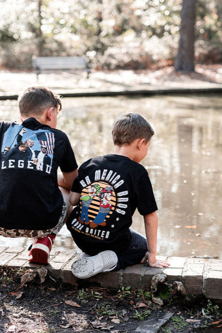 Two young boys sit on a ponds edge in retro tees. The boy on the left wears a black Youth Super Merica Bros T-shirt by Born United Private Label. Theyre perched on a stone border, surrounded by trees and a bench, capturing the carefree essence of youth.