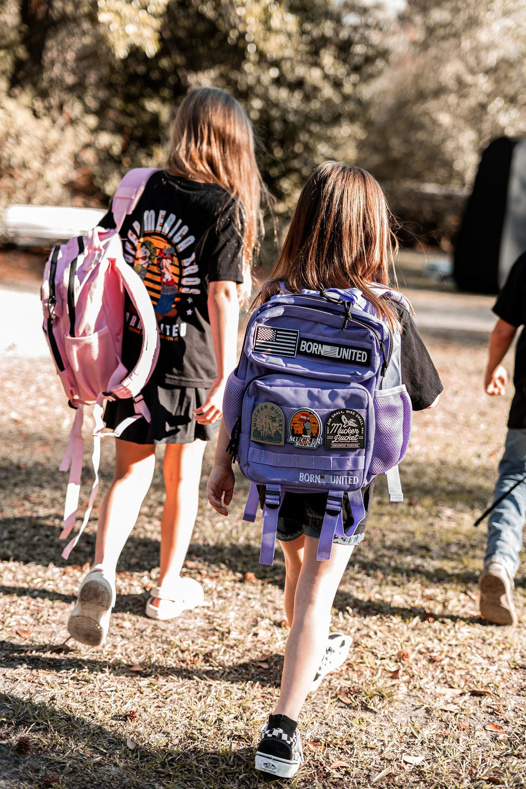 Two children walk on a grassy path enveloped by trees. The child on the left wears a pink backpack, while the one on the right carries a Born United Private Label Imperial Purple Tac-Pac - 15L with vibrant patches, embracing a playful and adventurous journey.
