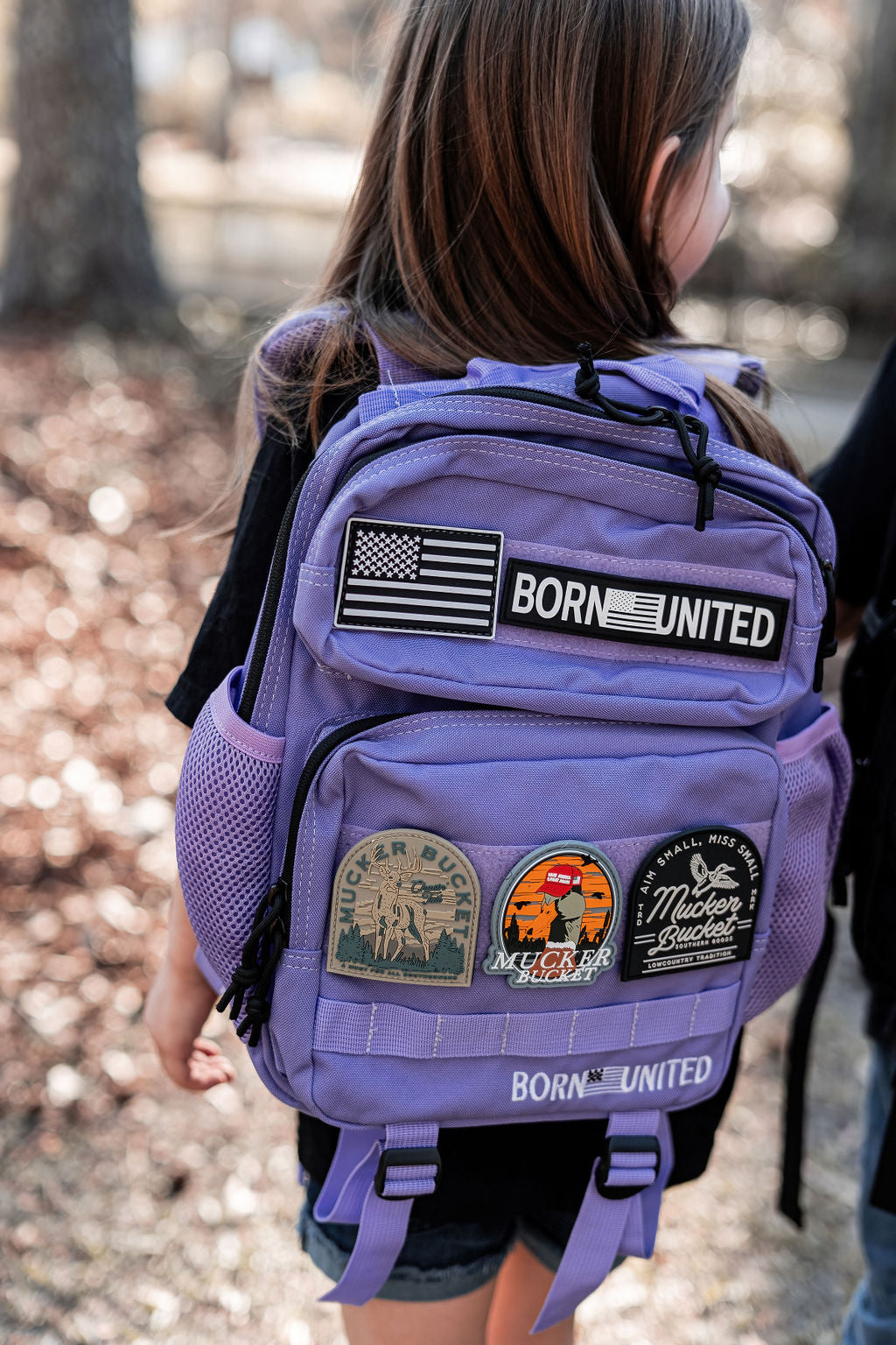A child with long brown hair sports a black shirt and an Imperial Purple Tac-Pac - 15L backpack by Born United Private Label. The backpack, featuring patches like an American flag and BORN UNITED, stands out against the softly blurred tree-filled background.
