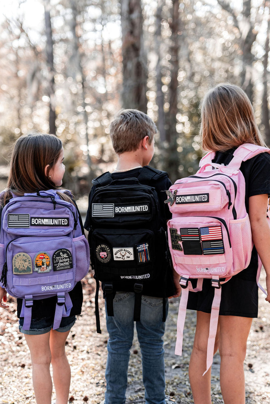 Three children outdoors, backs to the camera, wear backpacks with patches. The shades include purple, pink, and the Midnight Black Premium Tac-Pac - 15L by Born United Private Label. Trees and diffused sunlight softly illuminate the background.