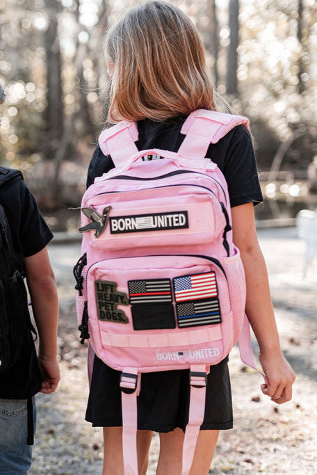 A girl sporting a Born United Private Label Pink Mist Premium Tac-Pac - 15L decorated with patches, including American flags and Lift Heavy Pet Dogs, stands outdoors on a sunny day, surrounded by a fresh breeze and next to another partially visible person.
