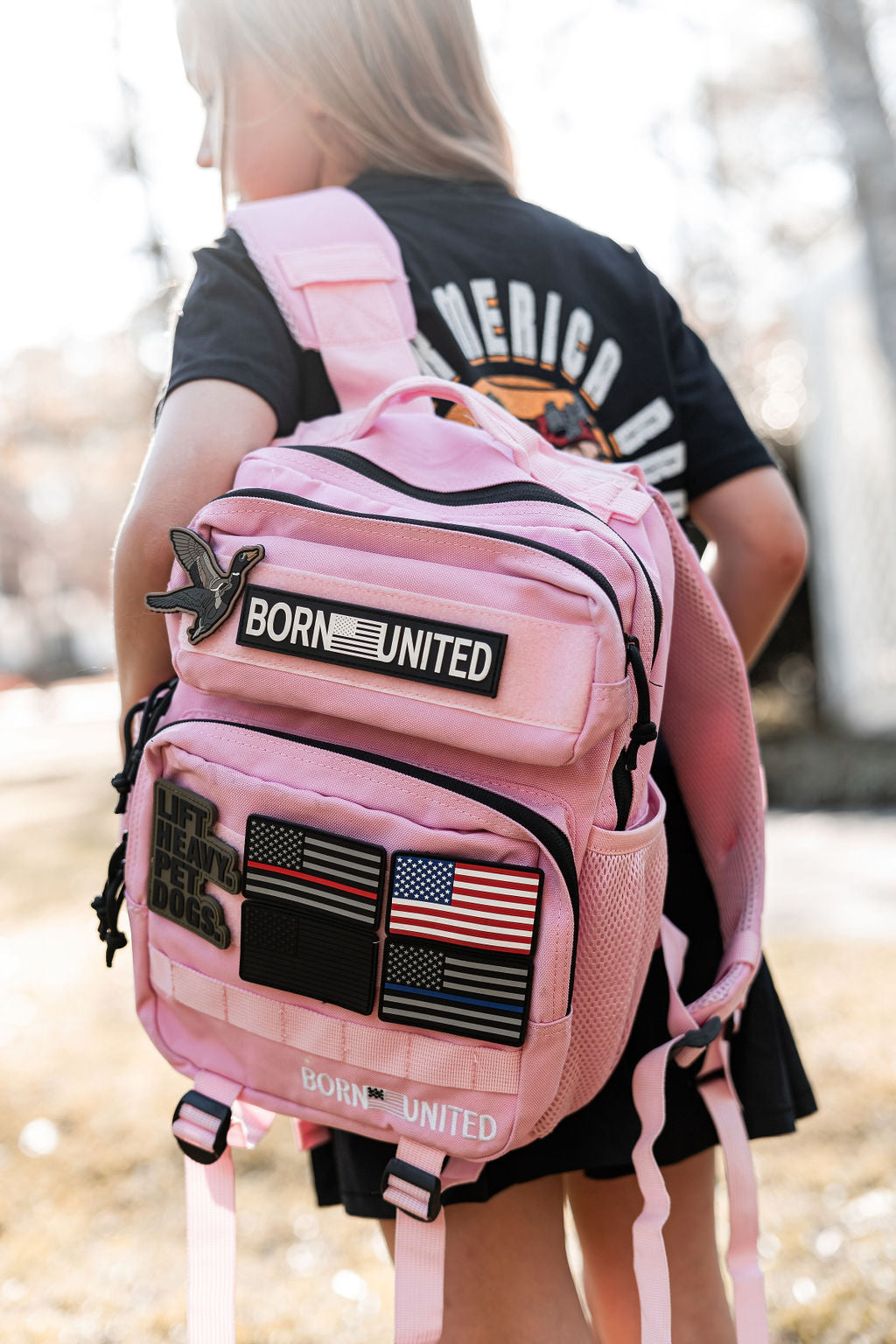 A person wearing a black shirt carries a Pink Mist Premium Tac-Pac - 15L by Born United Private Label, kissed by sunlight with patches like two American flags and ones reading “Born United” and “Lift Heavy,” creating an aura of delicate pink mist.