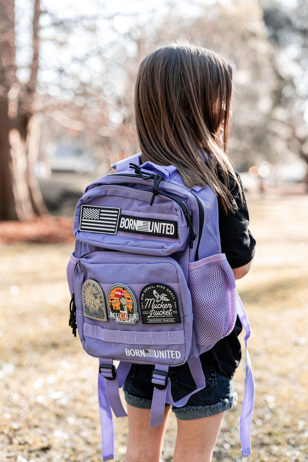 A child with long hair stands outdoors, wearing an Imperial Purple Tac-Pac - 15L featuring patches, including one with an American flag reading BORN UNITED, and a black shirt. Sunlight filters through the surrounding trees.
