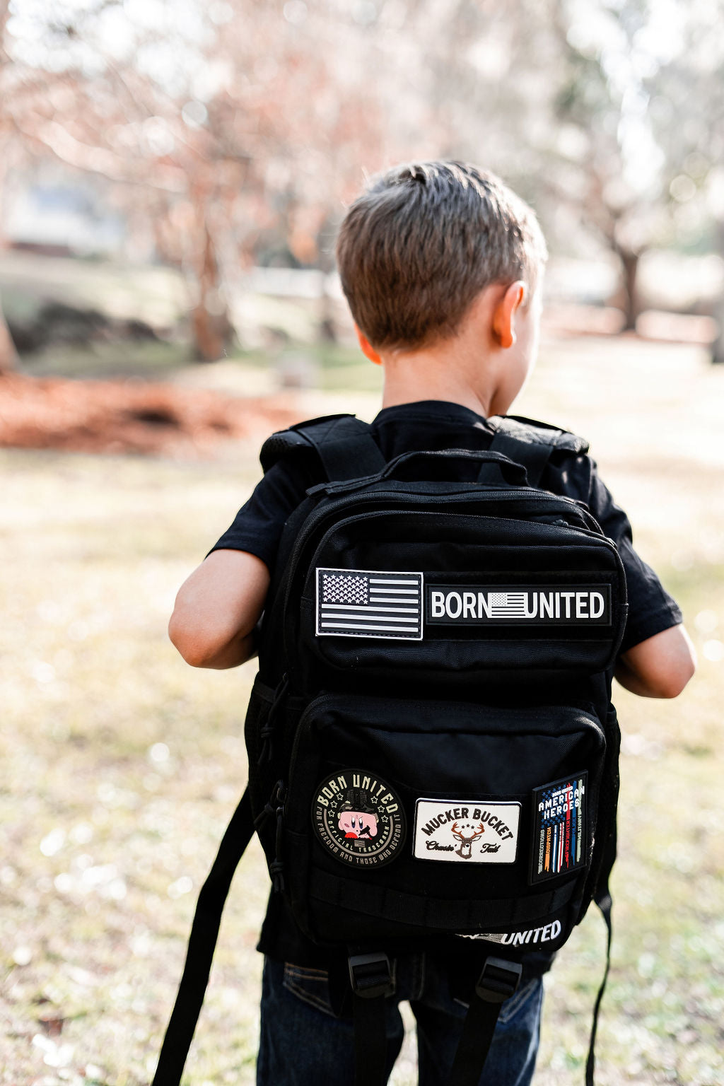 A young boy with brown hair stands outdoors, carrying a Midnight Black Premium Tac-Pac - 15L from Born United Private Label, featuring patches like an American flag and BORN UNITED, against a sunny park backdrop with lush grass and towering trees.