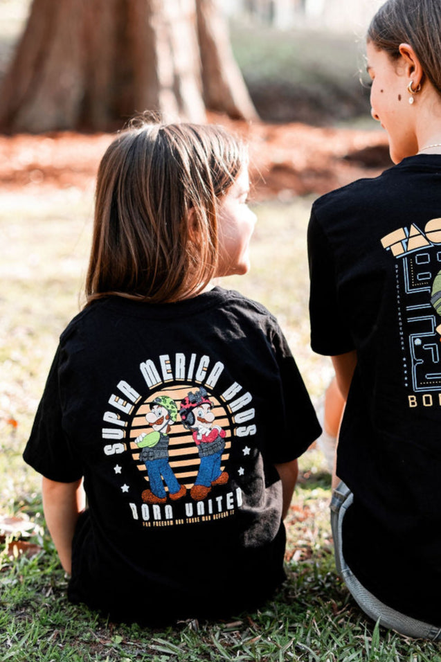 Two people sit on grass, seen from behind. The left person wears a Youth Super Merica Bros T-shirt by Born United Private Label, featuring cartoon characters and SUPER MERICA BROS and BORN UNITED. It evokes a vintage arcade game vibe amidst trees and leaves.