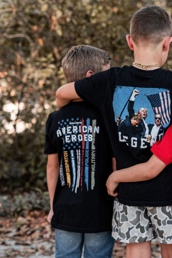 Two children outdoors, wearing youth-sized black T-shirts by Born United Private Label, stand with their backs to the camera. The left shirt says American Heroes, celebrating everyday heroes, and the right depicts a patriot-in-training with an American flag and a man.
