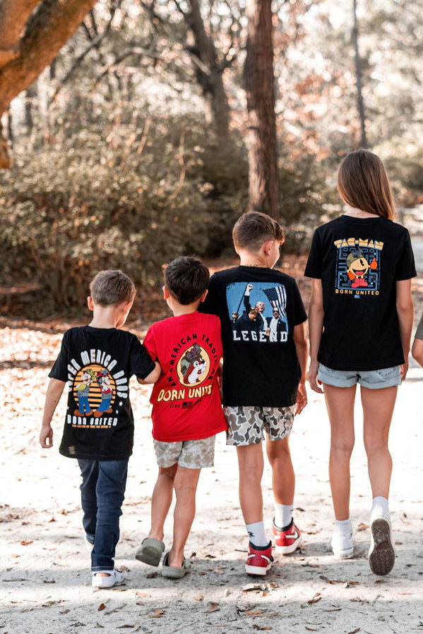On a sunny day, four kids walk outdoors wearing Youth Super Merica Bros tees from Born United Private Label in sizes XS-XL. The shirts feature colorful graphics reminiscent of vintage arcade games on the back as they stroll down a sandy path surrounded by trees and scattered leaves.