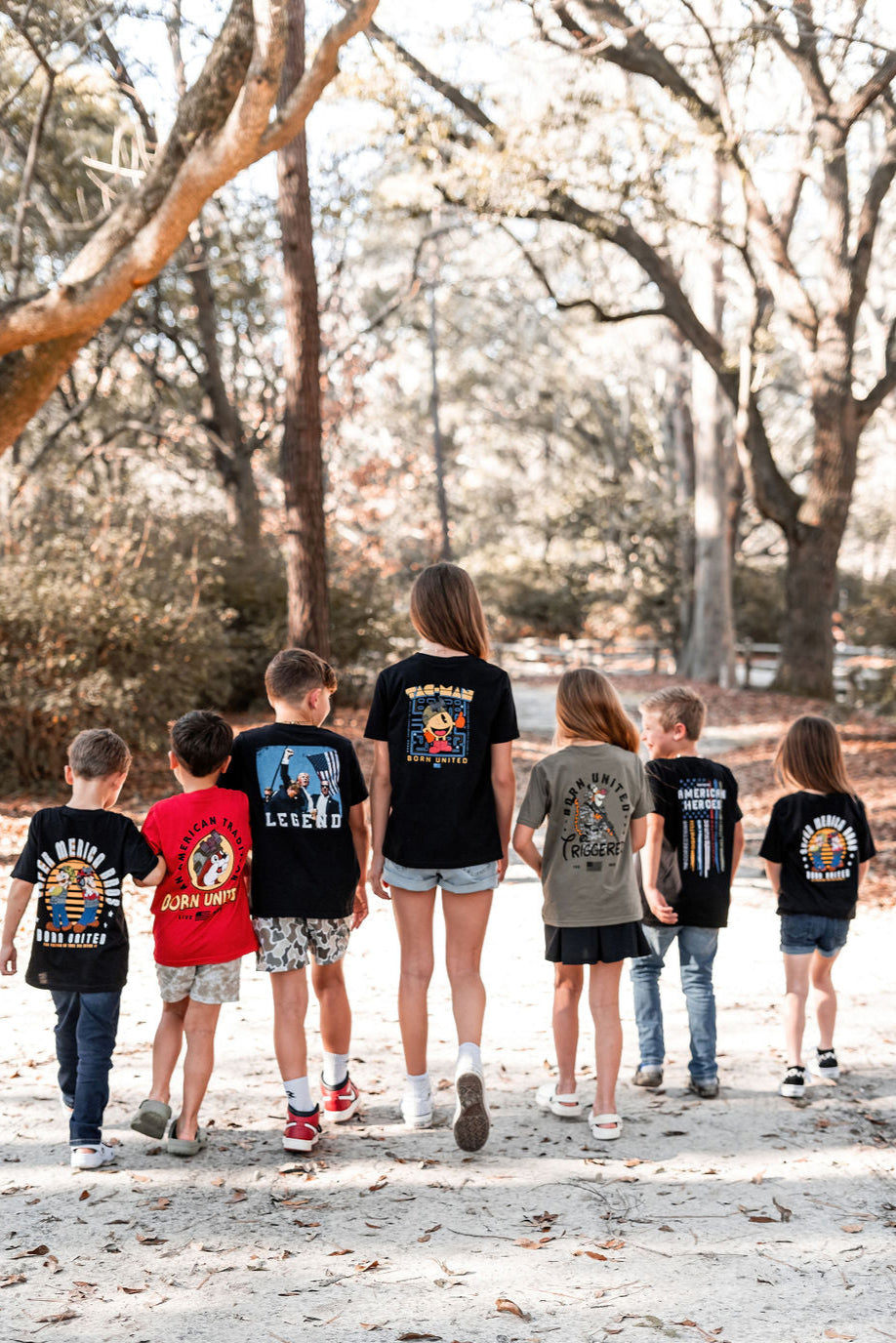 Seven children, resembling everyday heroes, walk along a sunlit forest path. They wear Youth American Heroes graphic t-shirts and shorts from Born United Private Label, surrounded by sparsely-branched trees suggesting fall or early spring.