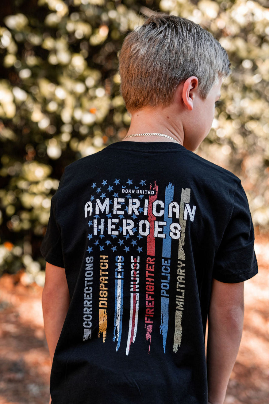A boy outdoors in a black Youth American Heroes T-shirt by Born United Private Label proudly displays the phrase American Heroes. The vertical stripes symbolize everyday heroes, from Corrections to Military services. With tree foliage behind, its perfect for the patriot-in-training.