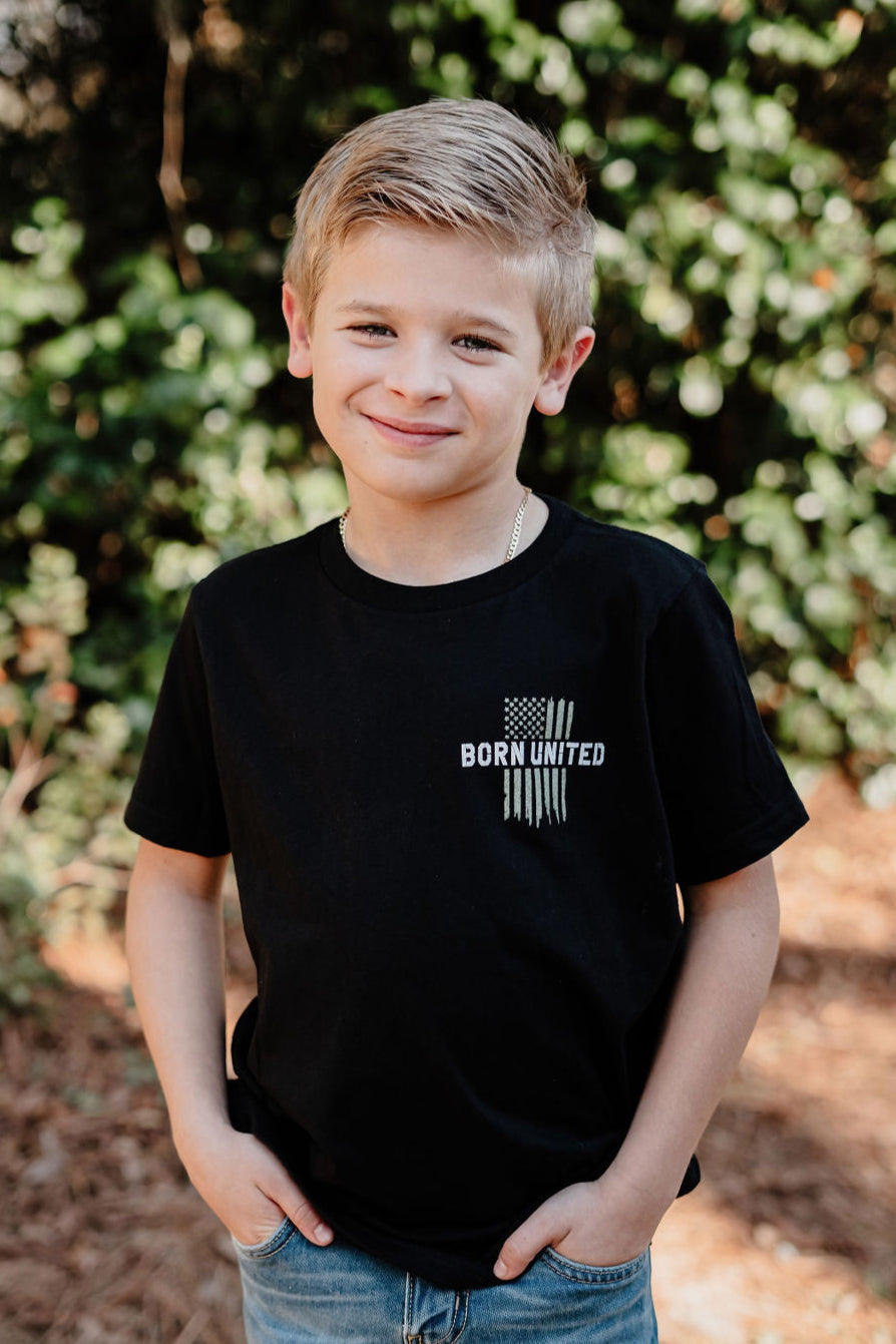 A boy with short blond hair stands outside, smiling in a black T-shirt by Born United Private Label. The shirt reads Youth American Heroes with a striped design. His hands rest casually in his pockets, embodying the spirit of youthful everyday heroes against lush greenery.