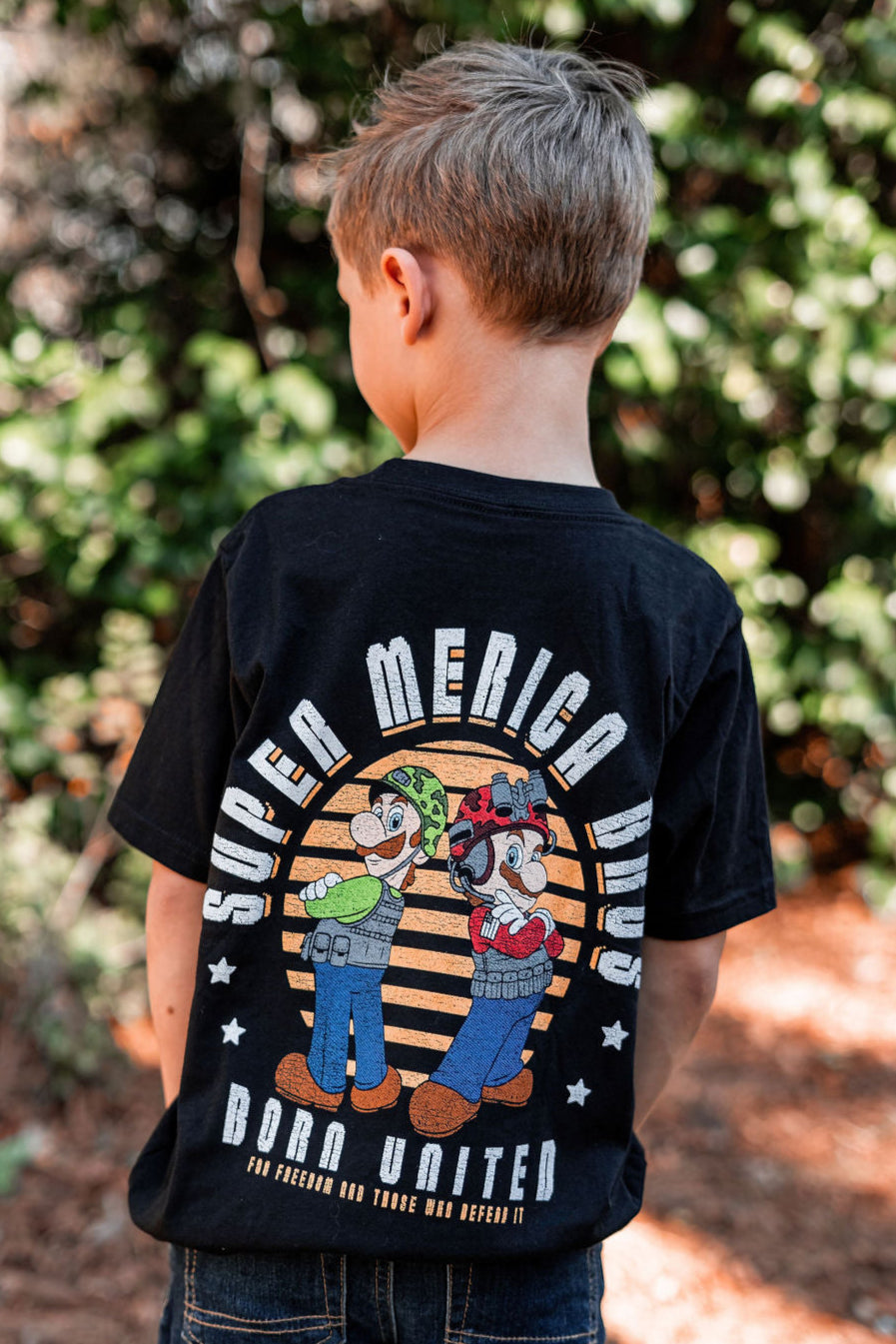 A young boy, back to the camera, stands outdoors in greenery wearing a black retro Youth Super Merica Bros tee by Born United Private Label. It features cartoon characters and Super America Bros Born United on a circular striped design. Available in youth sizes XS-XL.
