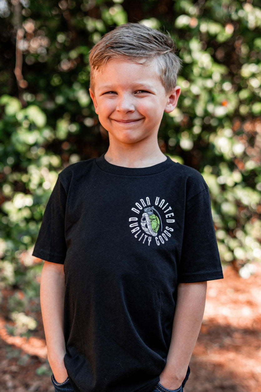 A young boy with short light brown hair stands outdoors, smiling with hands in his pockets. Hes wearing a Born United Private Label Youth Super Merica Bros retro T-shirt featuring a vintage arcade-style circular logo, against a backdrop of blurred greenery and dappled sunlight.