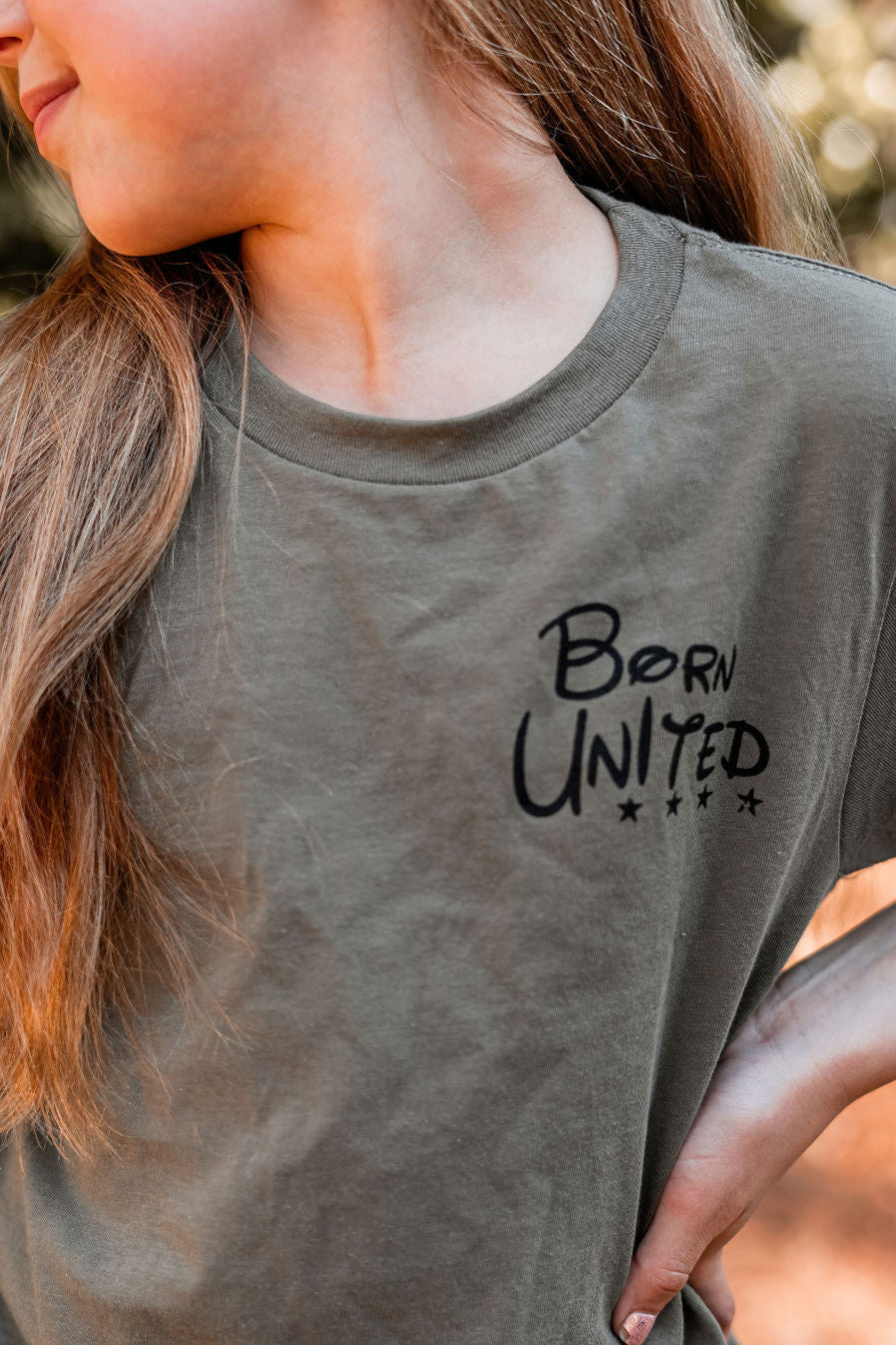 With long brown hair, a person shines against a blurred backdrop in the olive green Youth Triggered tee by Born United Private Label. Slightly turned to the right, they embody a patriot-in-training with quiet confidence.
