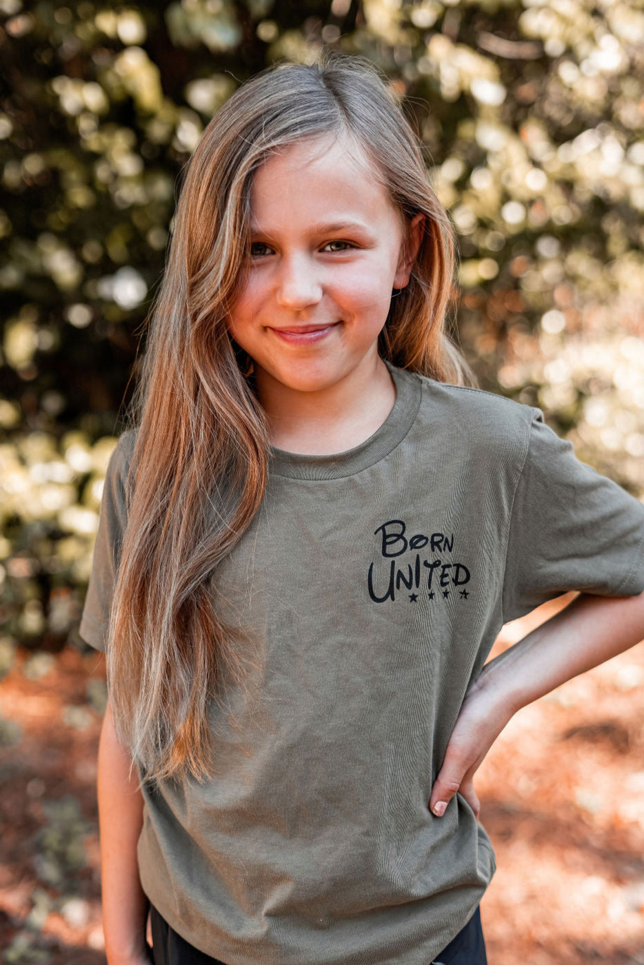 A young girl with long hair stands outdoors, smiling confidently at the camera. Shes wearing a green Youth Triggered tee from Born United Private Label, exuding patriotism in training. The soft-focus greenery in the background suggests a natural setting.