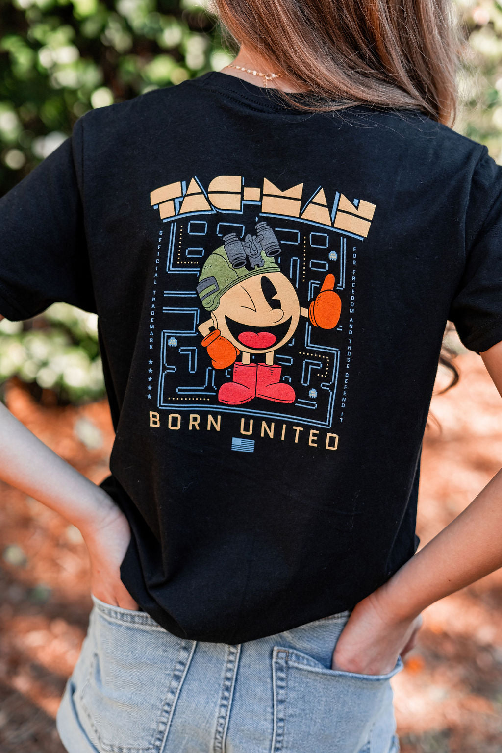 A patriot-in-training sports a black Youth Tac-Man T-shirt with a vintage arcade character. Born United is printed on the back as they stand outdoors, hands on hips, exuding confidence.