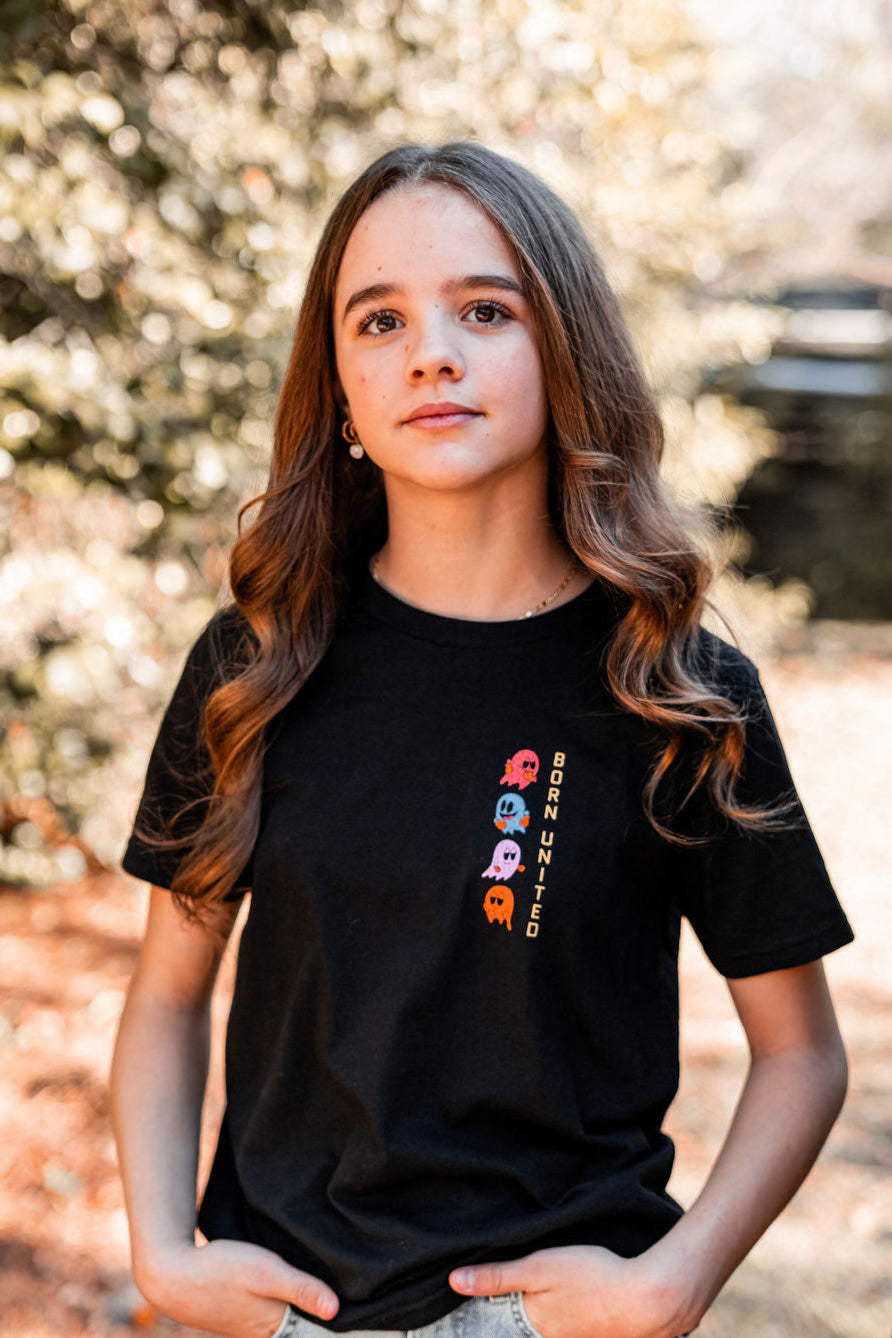 A young person with long brown hair stands outside in a black Youth Tac-Man T-shirt by Born United Private Label, featuring colorful arcade-style ghost designs and BORN UNITED. Blurred greenery and sunlight create the background. Available in youth sizes XS-XL.