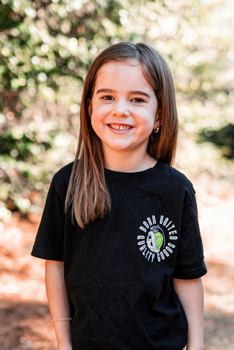 A young girl with long brown hair smiles at the camera, wearing a black Youth Super Merica Bros tee from Born United Private Label. The softly blurred greenery in the background hints at an outdoor setting.