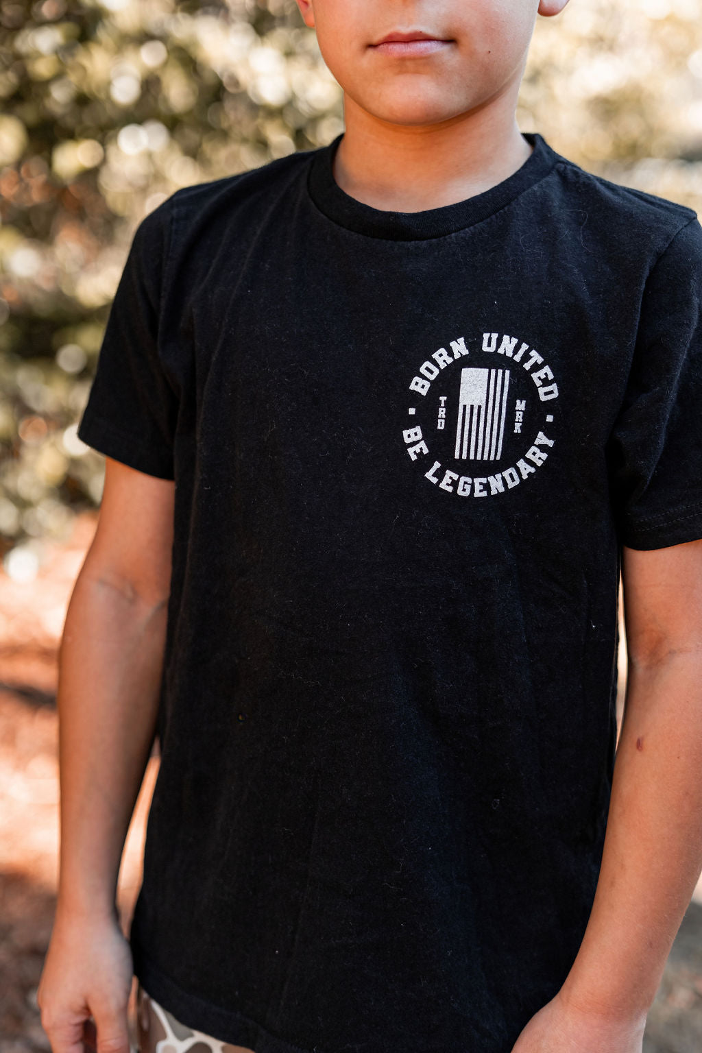 A child proudly wears a Youth Forever Legend T-shirt from Born United Private Label outdoors, with natural light filtering through blurred foliage in the background.