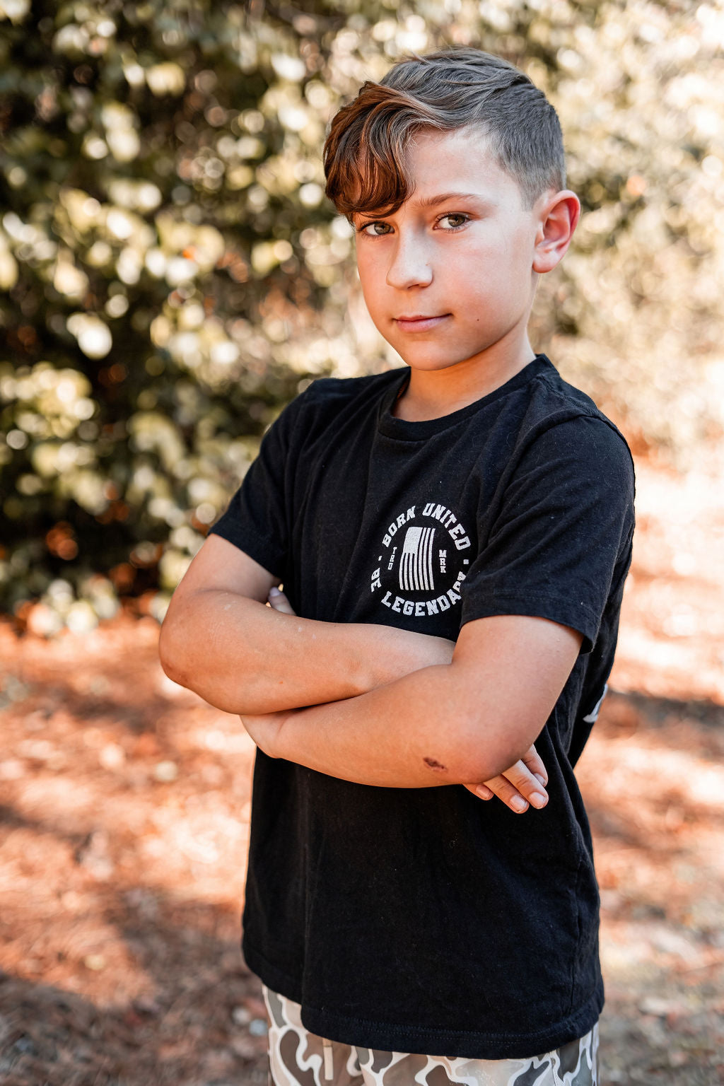 A young boy with short, styled hair stands confidently with arms crossed, wearing a Youth Forever Legend tee by Born United Private Label. His shorts are patterned. The background features blurred trees and sunlight, suggesting a natural setting.