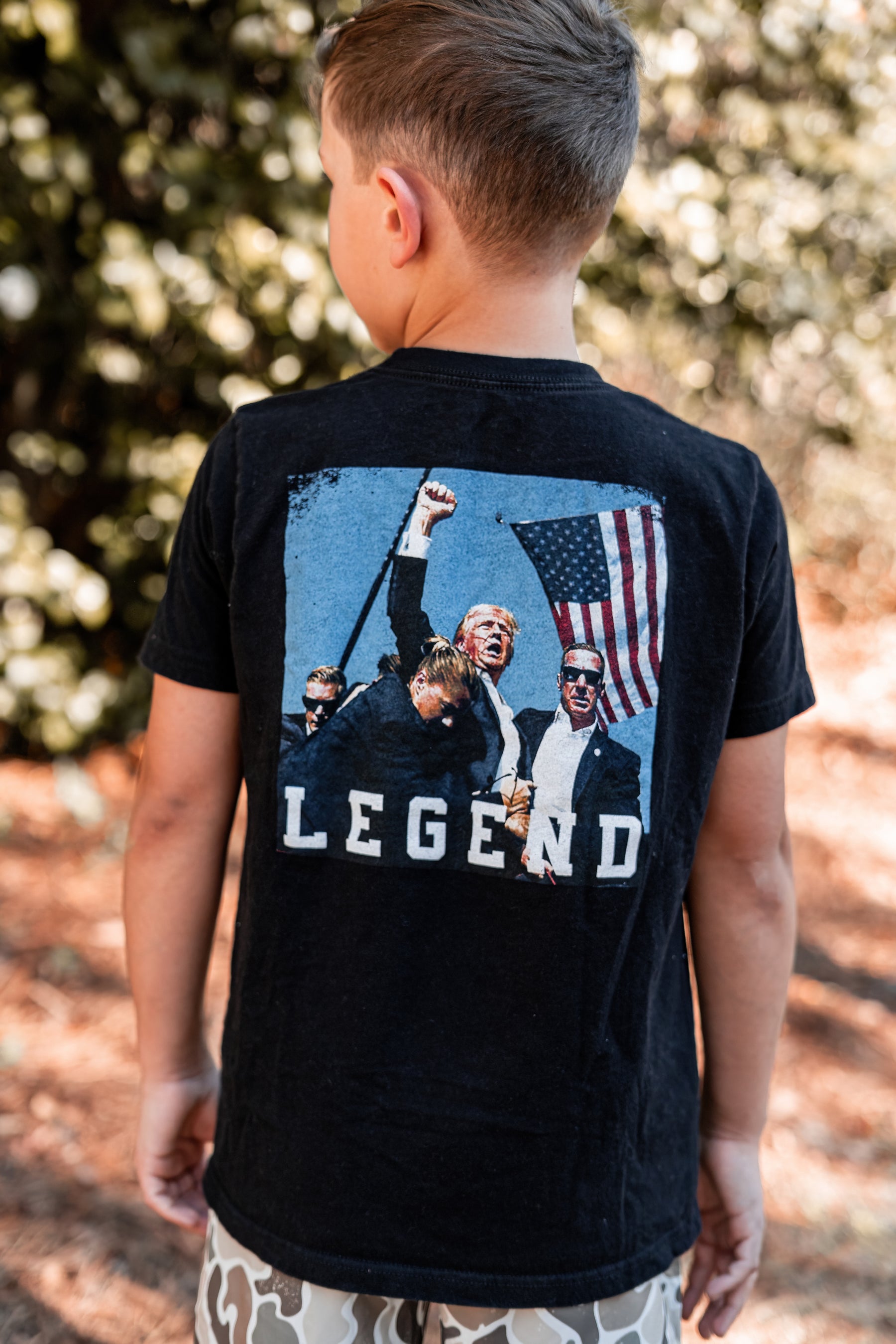A boy in a Youth Forever Legend tee from Born United Private Label, featuring four men with an American flag, stands outside against a backdrop of trees, exuding subtle patriot pride.