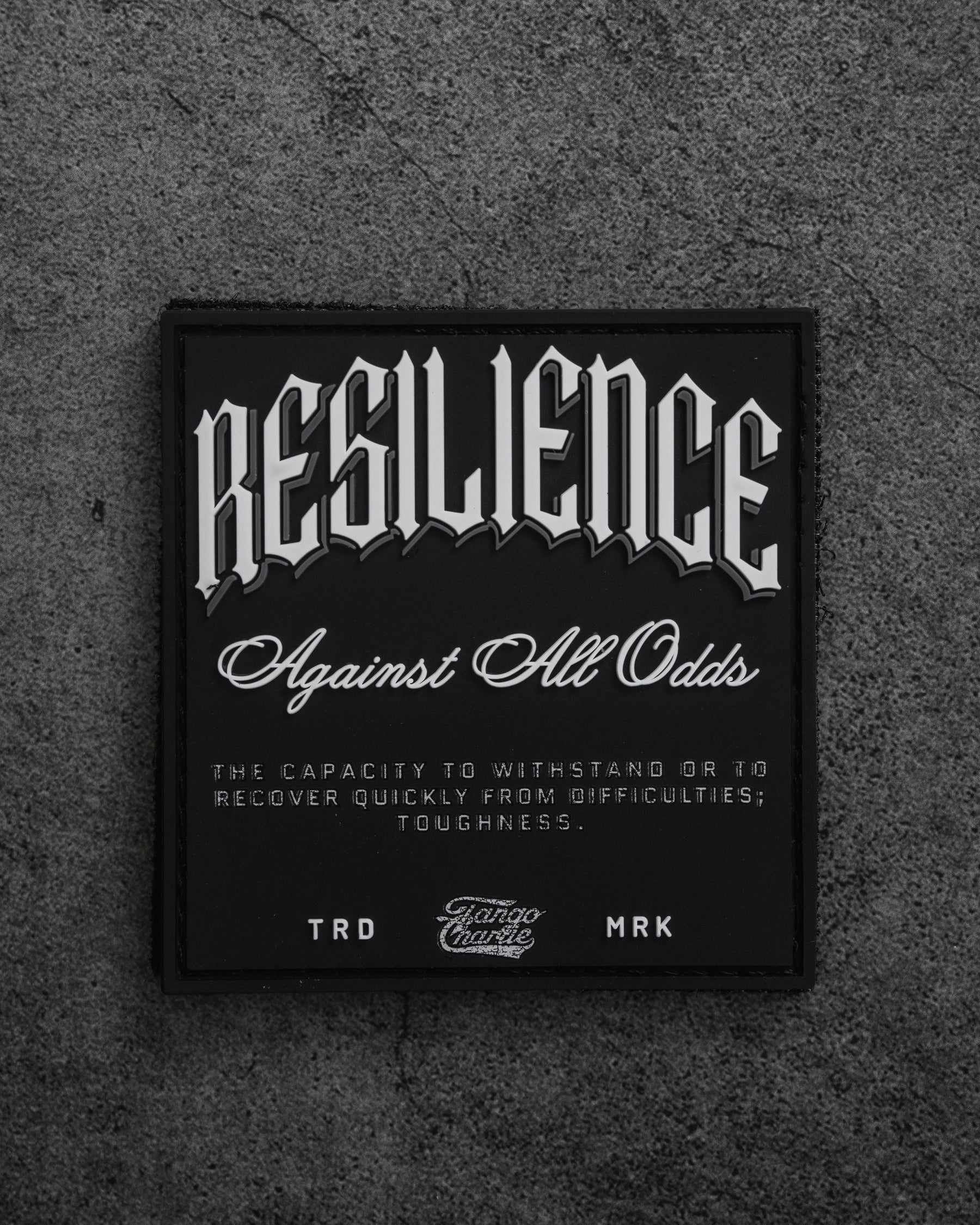 The Resilience-PVC Patch by Tango Charlie Apparel features bold RESILIENCE Against All Odds text and a definition on a black patch with a gray background, symbolizing toughness and the capacity to recover from difficulties.
