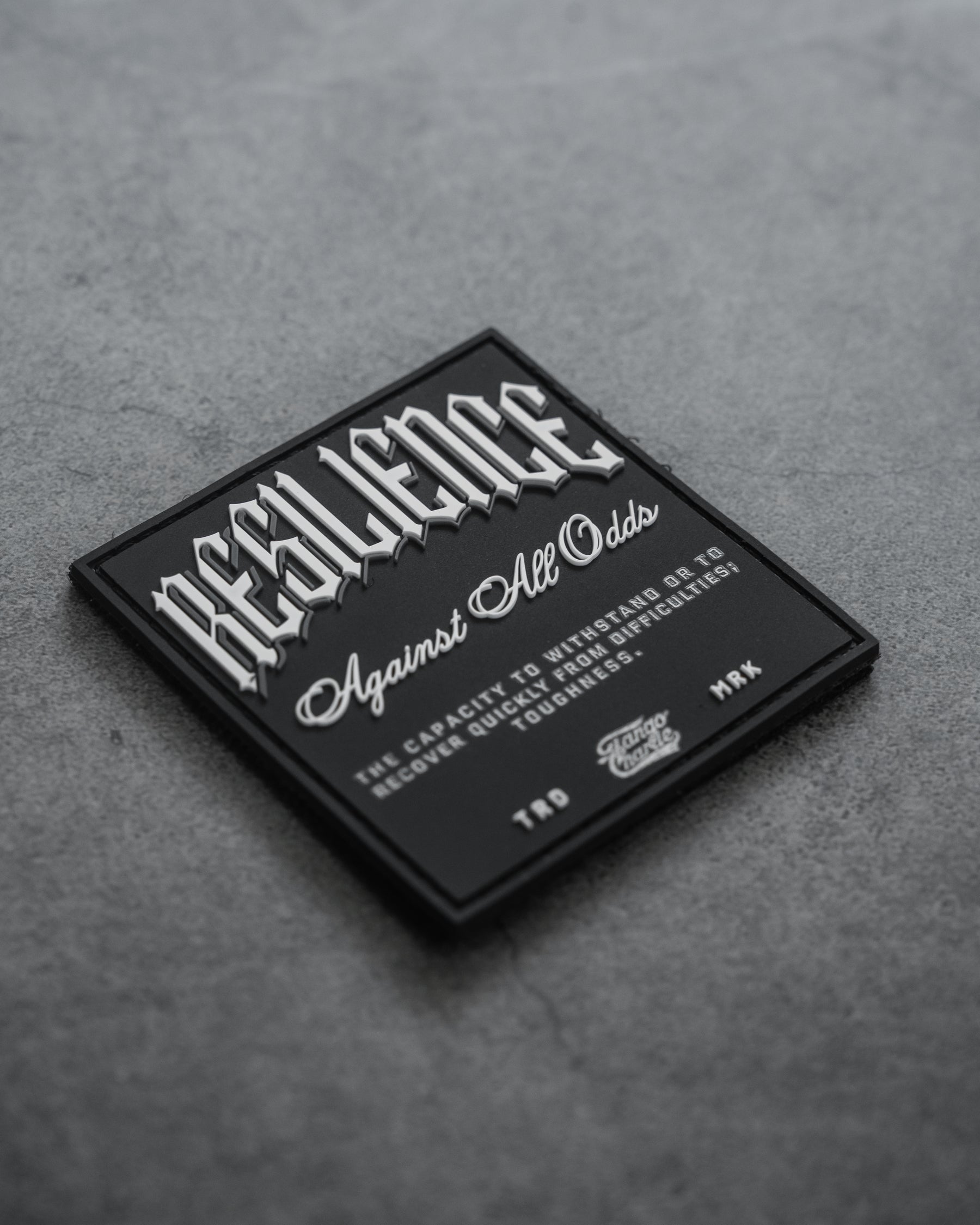 The Resilience-PVC Patch by Tango Charlie Apparel features white text, RESILIENCE Against All Odds: The capacity to withstand or to recover quickly from difficulties; toughness, on a black square patch displayed on a gray textured surface.