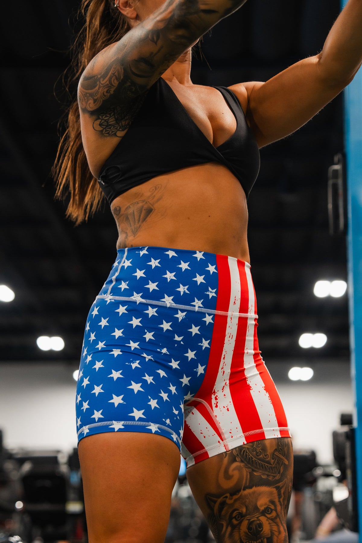 A woman with tattoos and long hair works out in a gym, wearing Born United Private Labels Womens High Waisted Seamless Front Compression Training Shorts | Stars & Stripes. Gym equipment is visible in the background.