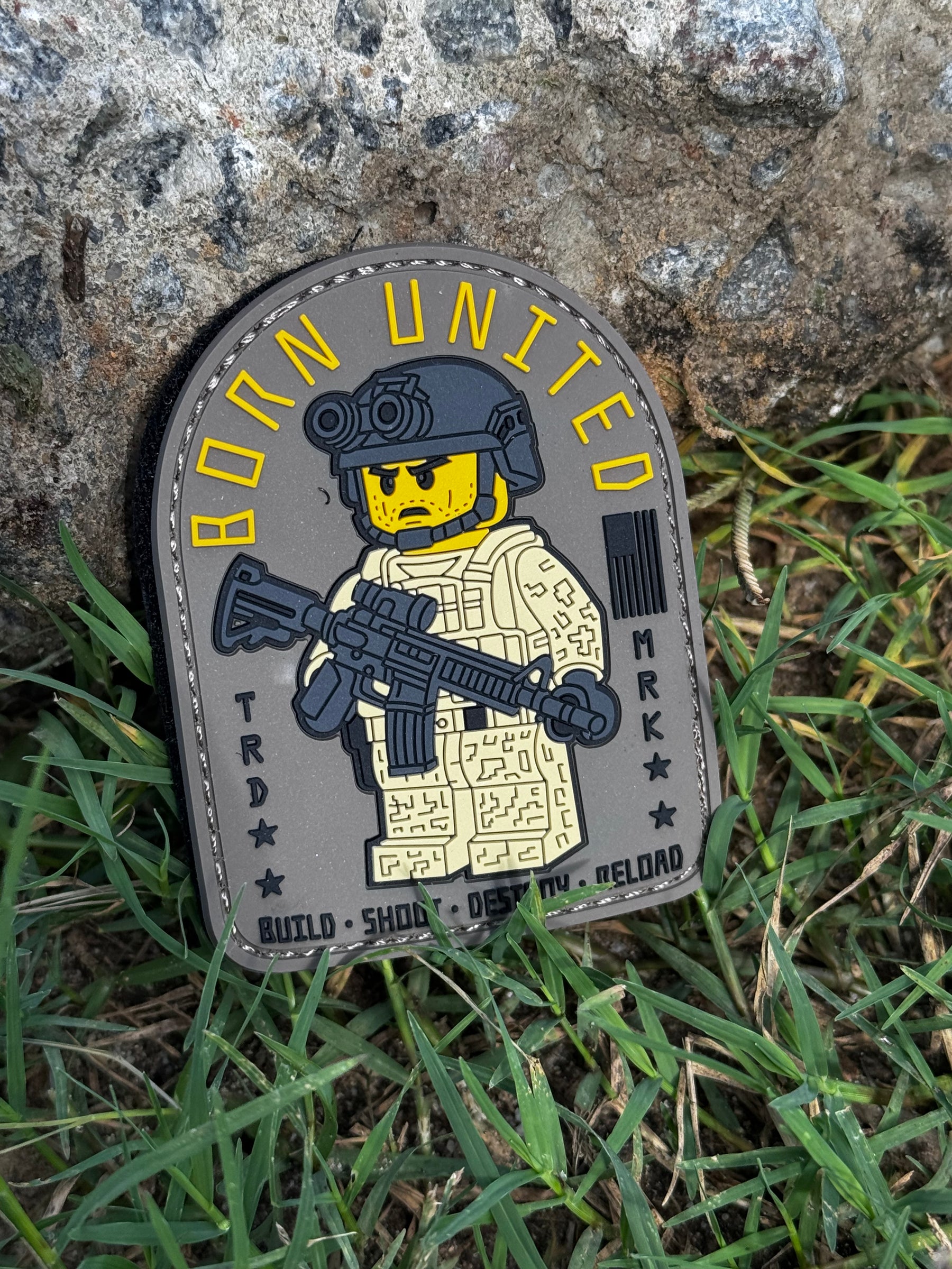 The Born United Private Label Brick Builder PVC Patch shows a LEGO-style soldier in tactical gear with a rifle under the words BORN UNITED, made from durable PVC and pictured propped on grass against a rock.
