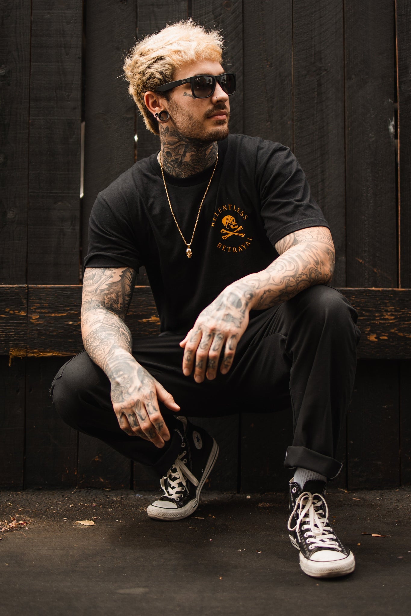 A tattooed man with blond hair and sunglasses crouches in front of a dark wooden fence. He wears a Relentless Betrayal Lost At Sea Black T-shirt made of fine jersey cotton, black pants, black Converse shoes, and a gold chain that enhances the edgy UNISEX style.