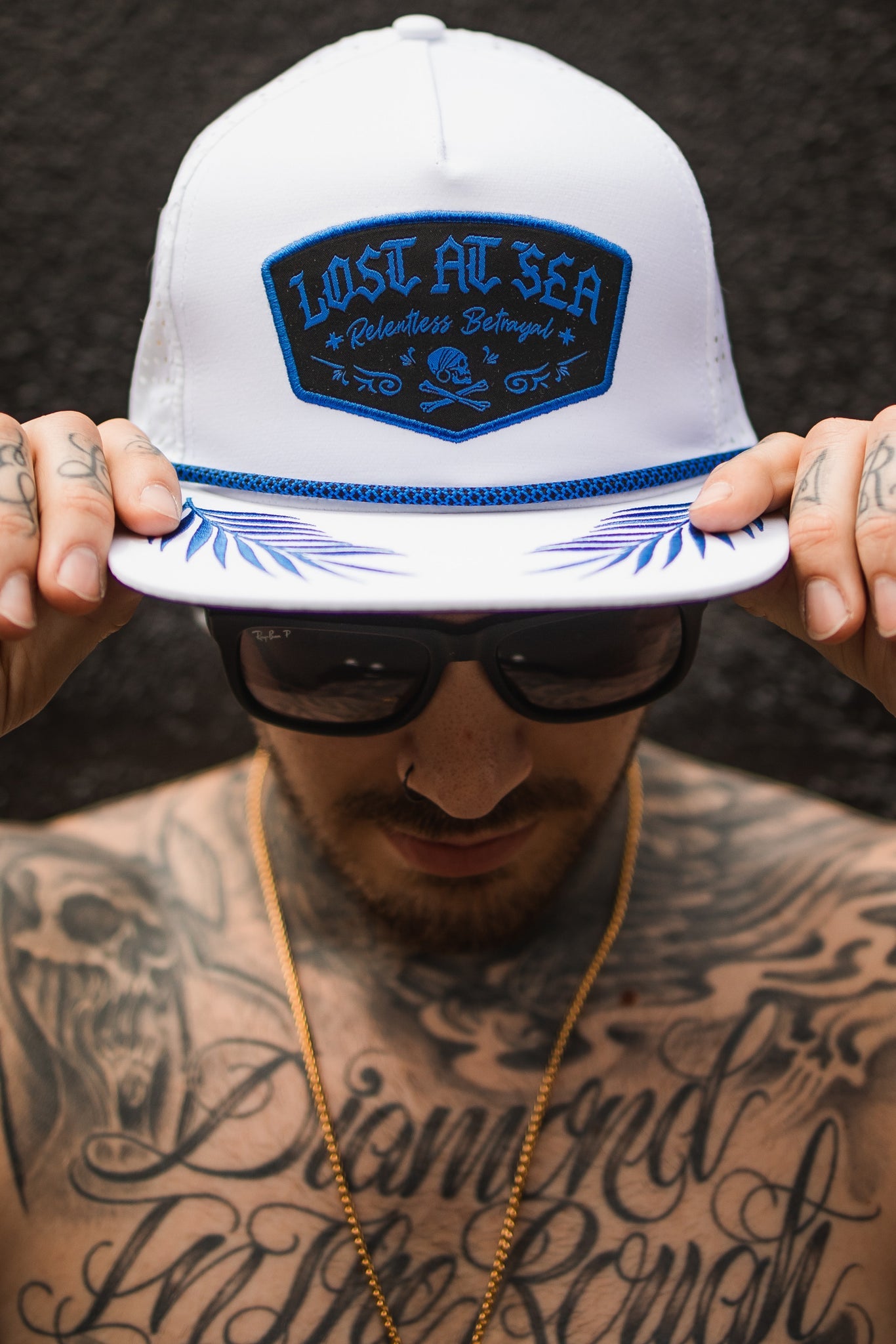 A tattooed individual holds the brim of a Relentless Betrayals Lost at Sea Whiteout Snapback cap. They are shirtless, wearing sunglasses and a gold chain against a dark, textured background.