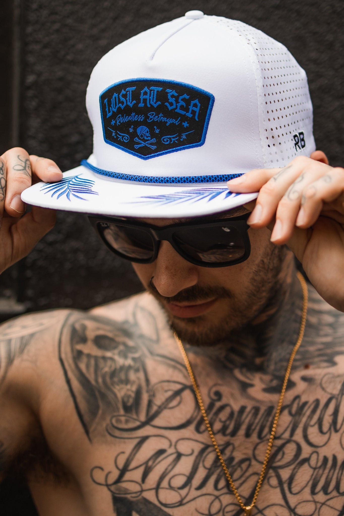A man adjusts the brim of his Relentless Betrayal Lost at Sea Whiteout Snapback, featuring a blue LOST AT SEA patch. Tattoos cover his chest and hands as one grips the cap, while a gold chain adorns his neck.