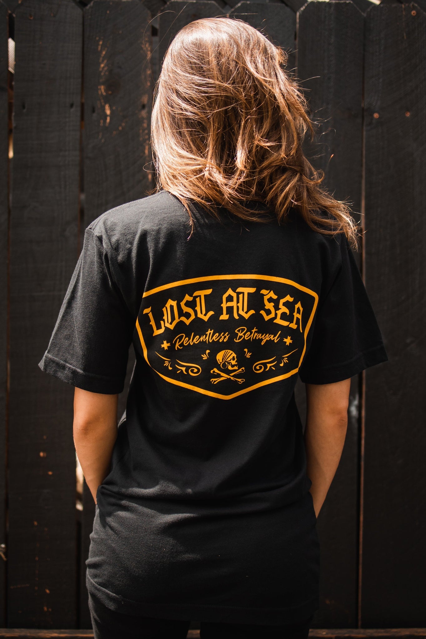 A person wears a Lost At Sea unisex black tee by Relentless Betrayal, featuring a yellow graphic with a skull motif. They stand with their back to us against a sunlit dark wooden fence.