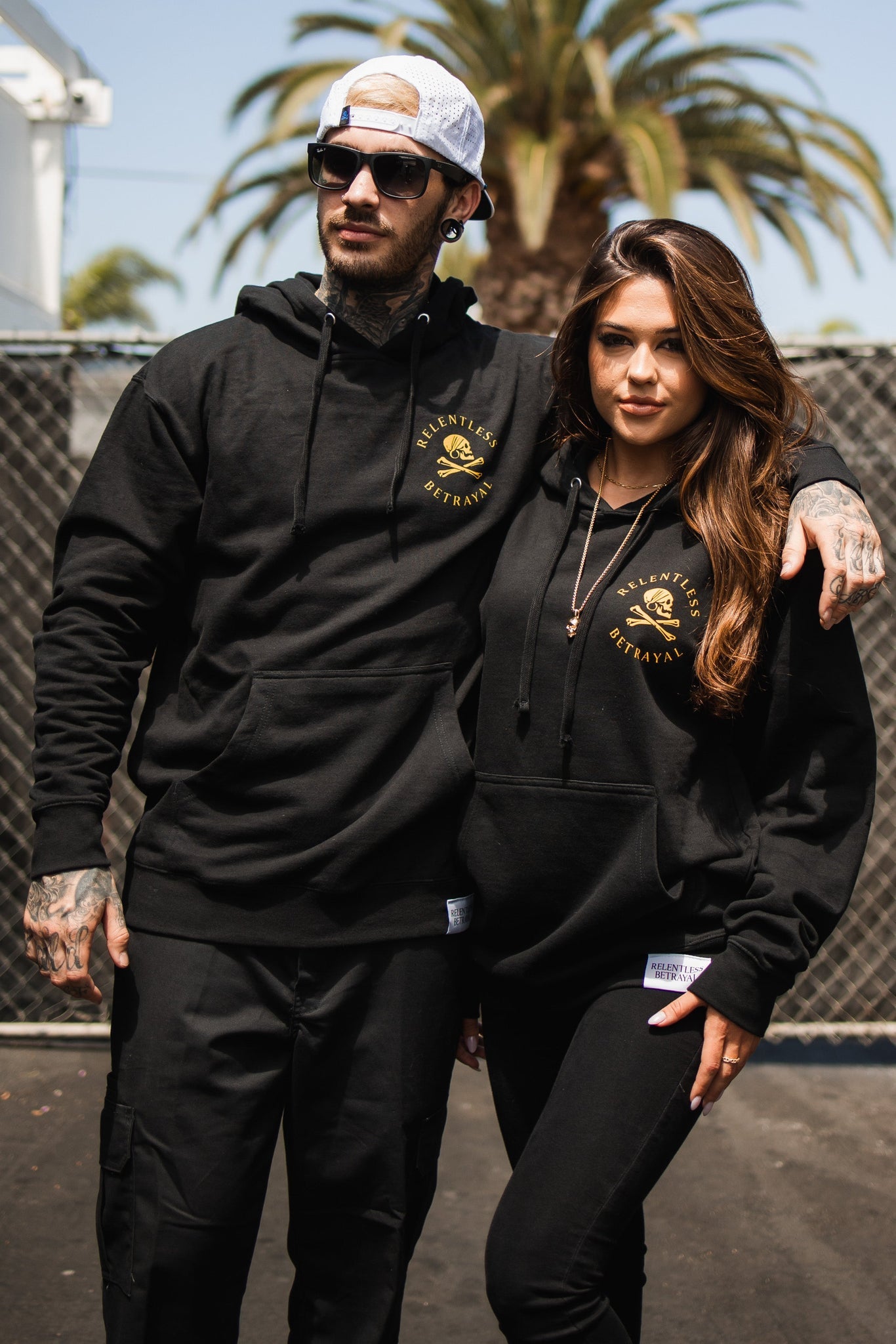 A man and woman pose confidently in unisex Lost at Sea black hoodies by Relentless Betrayal, featuring yellow logos. The man adds sunglasses and a backward snapback to his look. Standing on a paved surface with palm trees swaying behind, they effortlessly blend style and comfort.