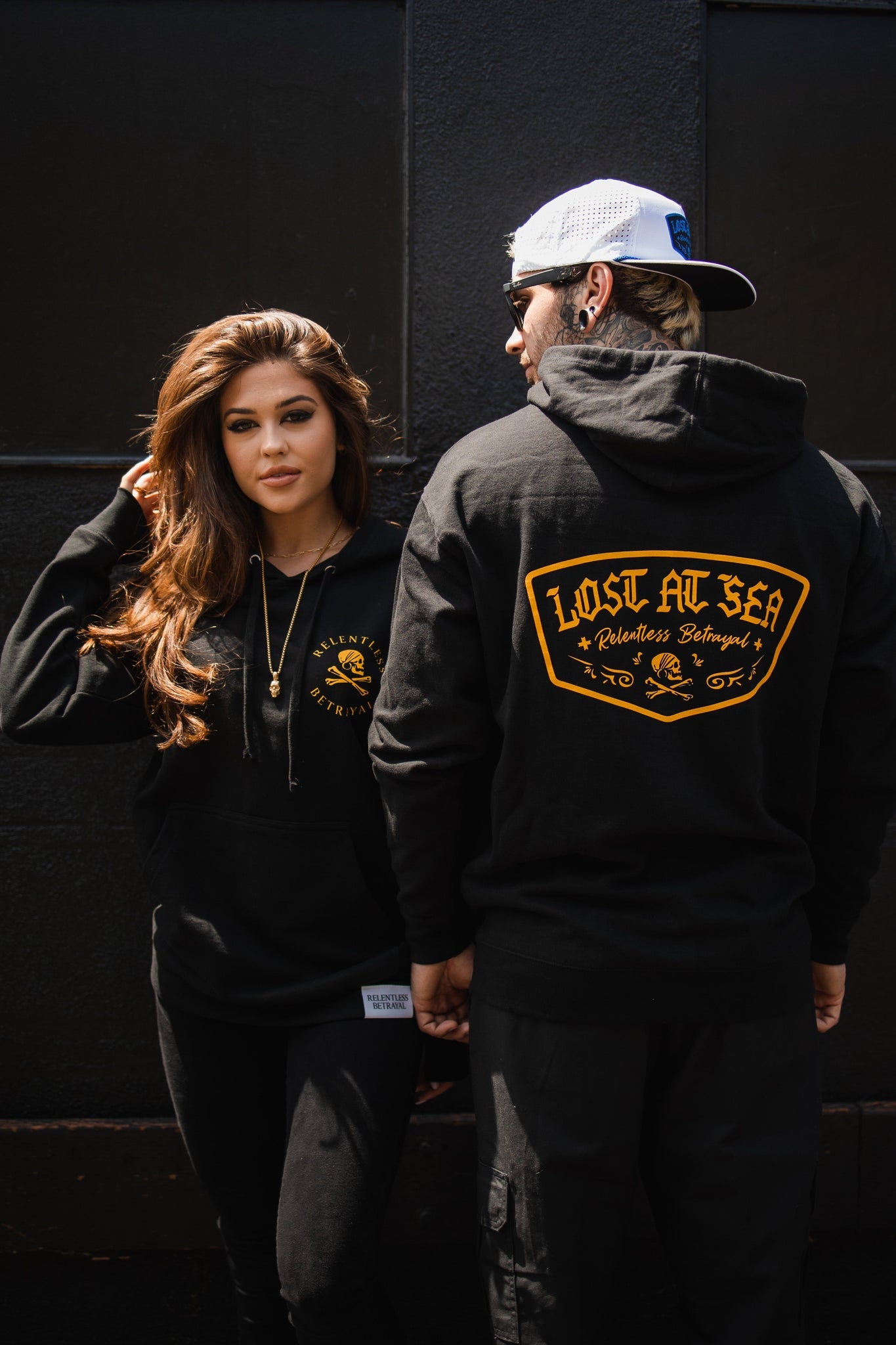 A woman and man in black hoodies from Relentless Betrayal stand against a dark wall. Hers, featuring a small logo, is made of a cotton/polyester blend, while his Lost at Sea Hoodie reads Lost At Sea on the back. Her long hair and his backward snapback cap complete their unisex style.