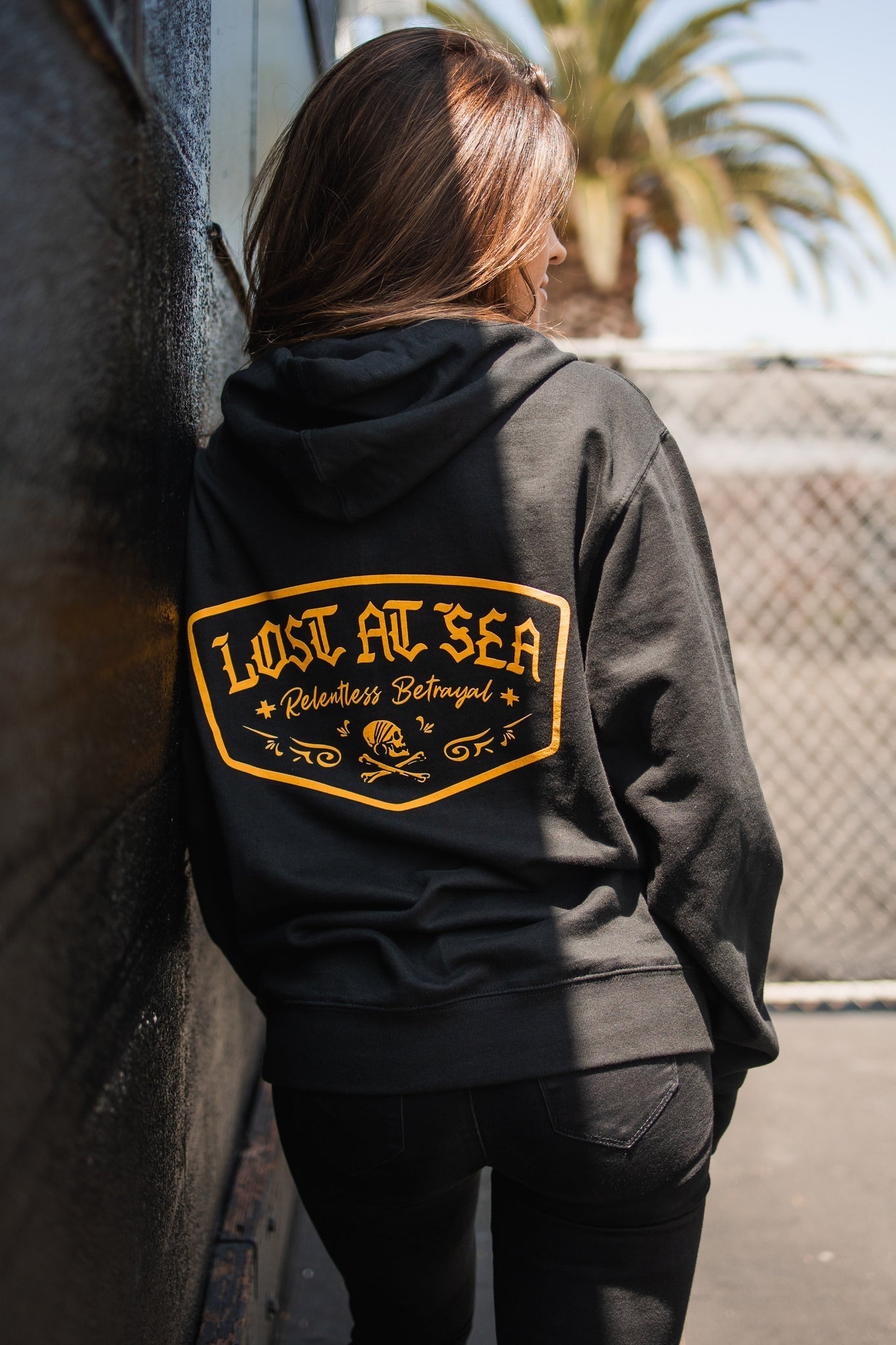A person with long hair in a black Lost at Sea Hoodie by Relentless Betrayal, featuring a cotton/polyester blend and a yellow design on the back, leans against a dark wall. In the background are a snapback, palm tree, and chain-link fence.