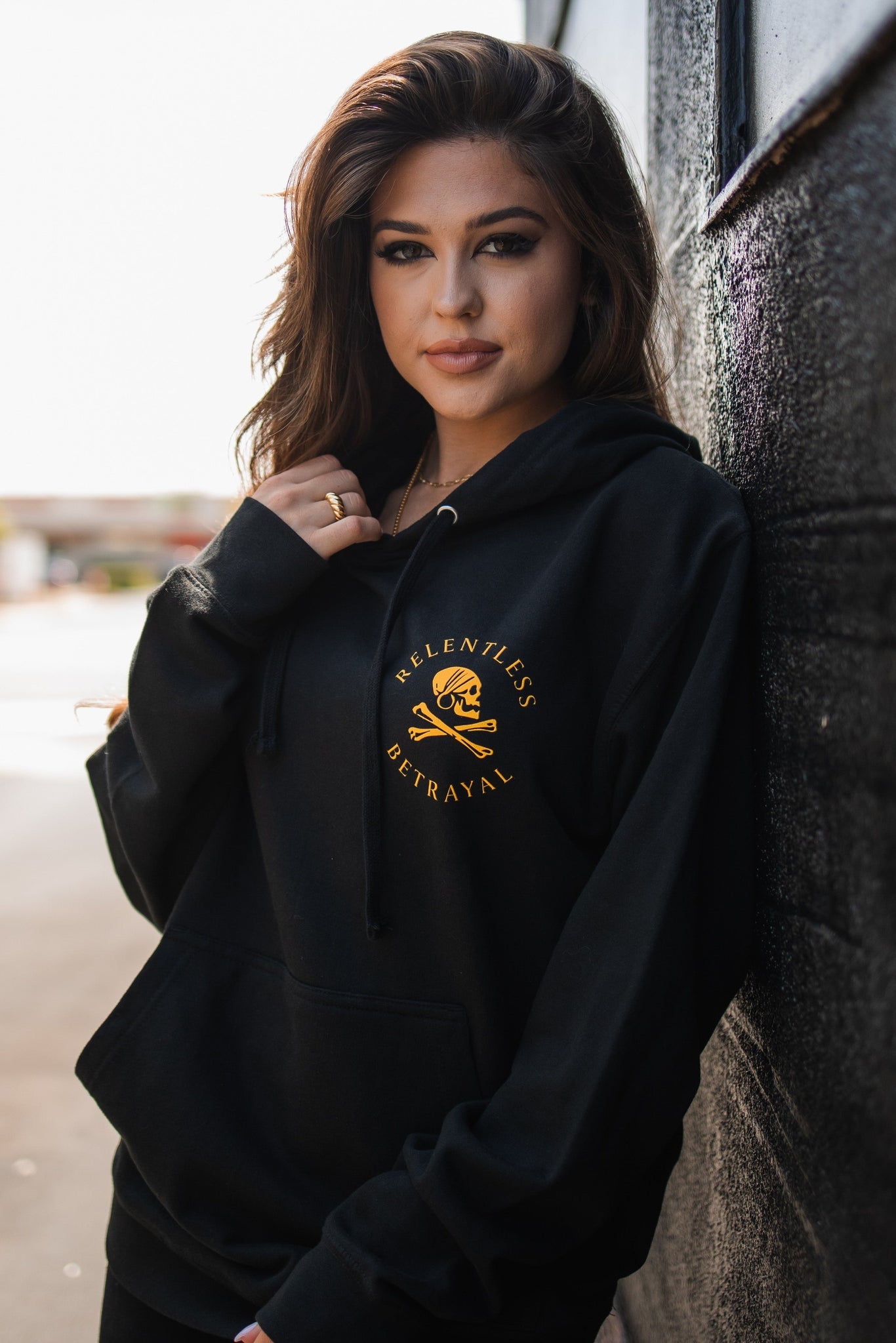 A woman with long hair is outdoors wearing a black Lost at Sea Hoodie by Relentless Betrayal. She leans against a wall, adjusting her unisex snapback while looking at the camera, sunlight emphasizing the soft cotton/polyester fabric.