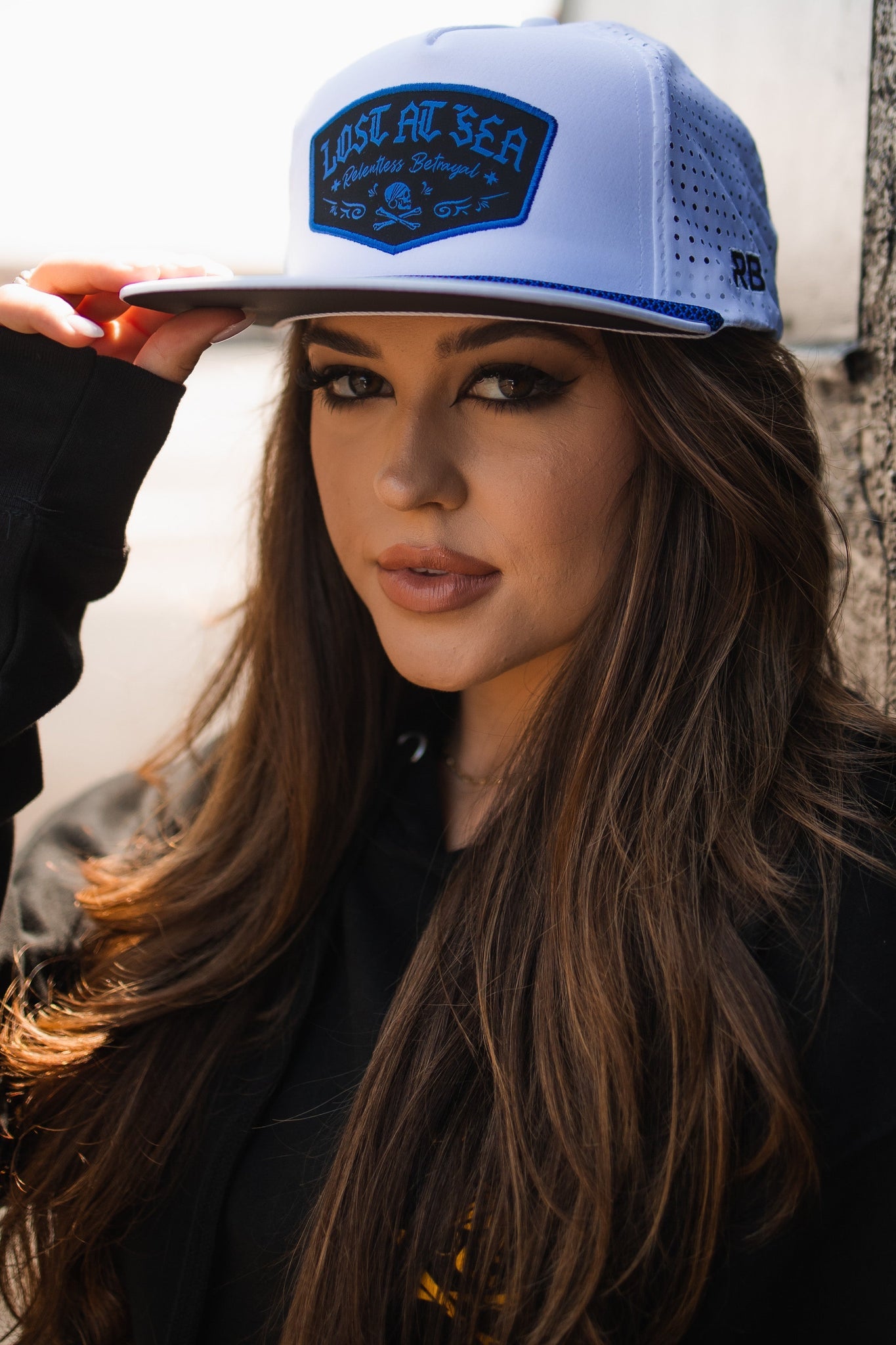A woman with long brown hair wears the Relentless Betrayal Lost at Sea Whiteout Snapback, a water-resistant white cap, along with a black hoodie, gazing confidently into the camera outdoors.