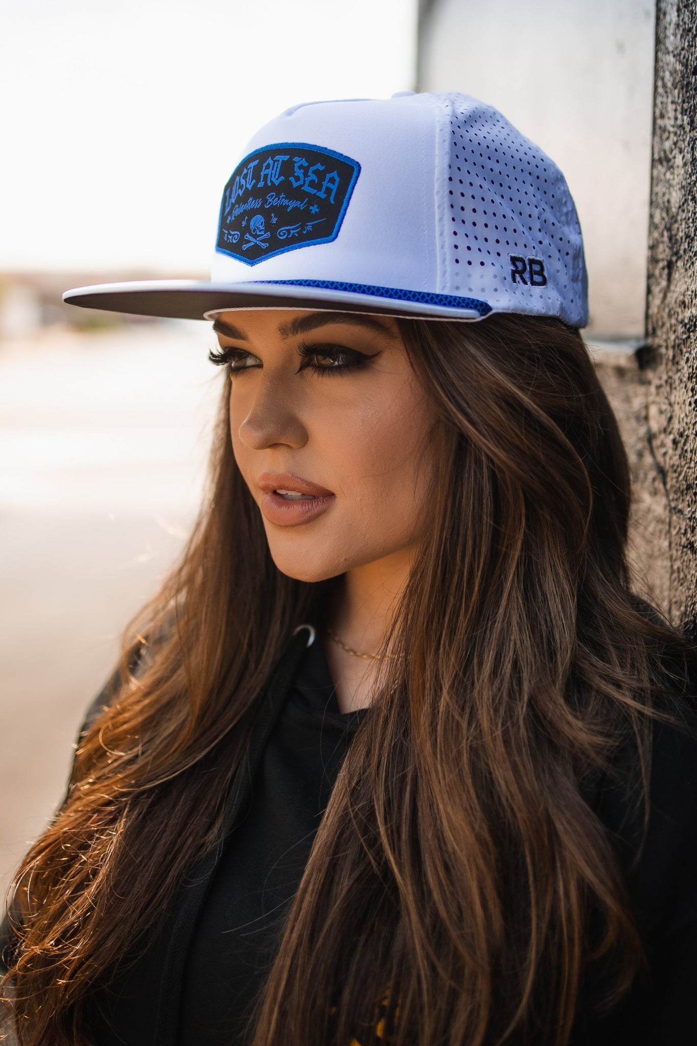 A woman with long brown hair wearing a Relentless Betrayal Lost at Sea Whiteout Snapback and a black hoodie gazes to the side. The water-resistant white cap features a blue patch with text, against a softly blurred background.