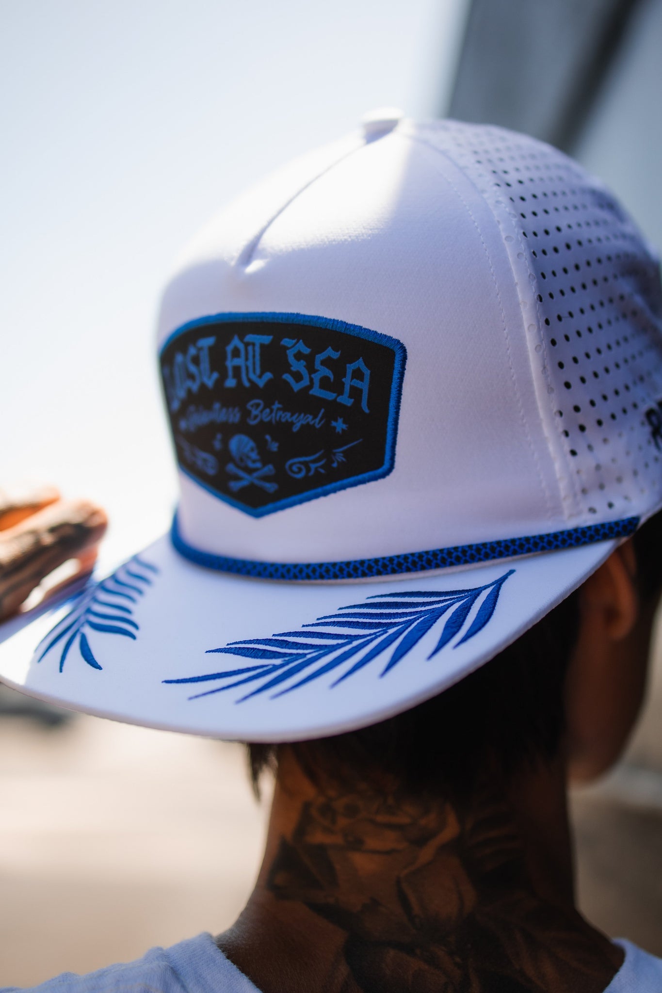 A person adjusts a Relentless Betrayal Lost at Sea Whiteout Snapback with palm frond designs on the brim, slightly tilted to show neck tattoos, against a blurred outdoor background.